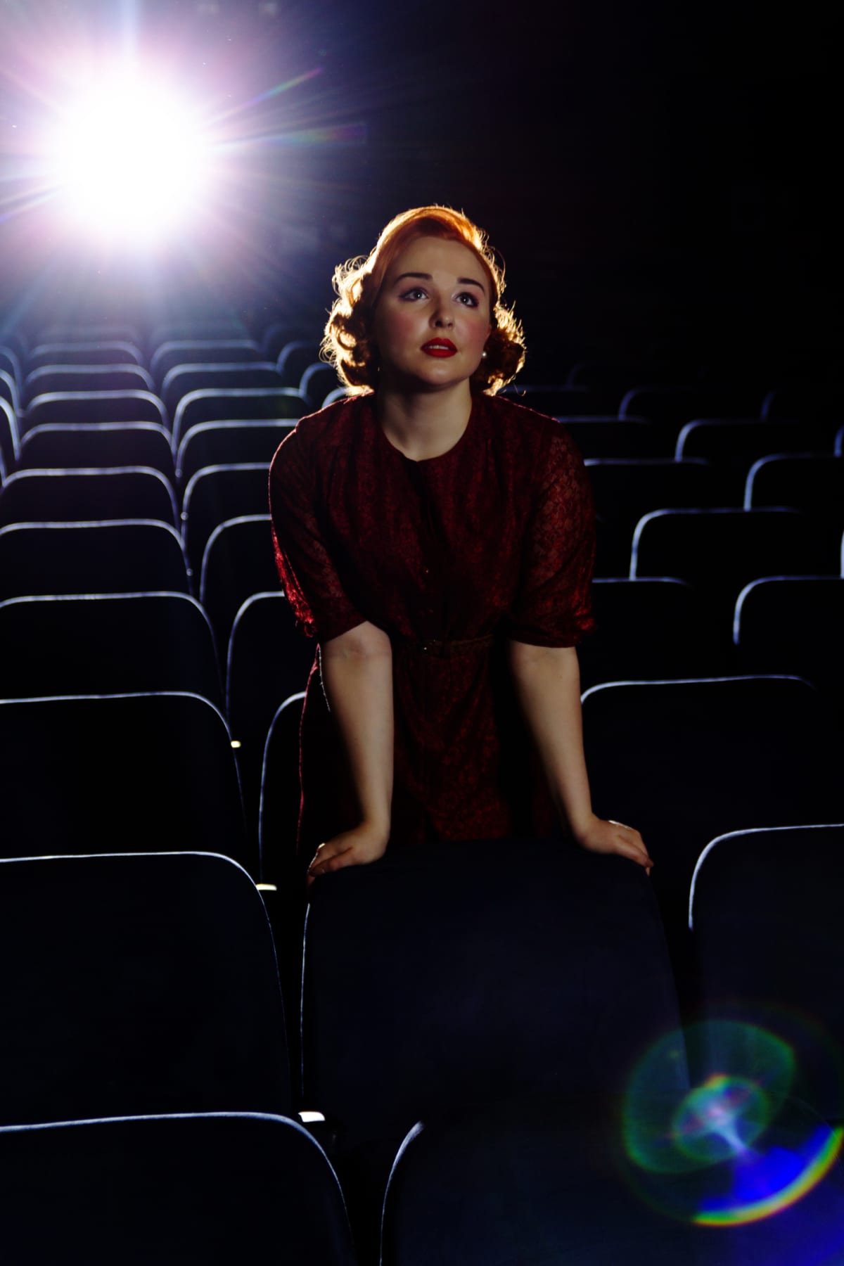 A young woman wearing vintage clothes stands in an empty cinema with the light of the projector behind her.
