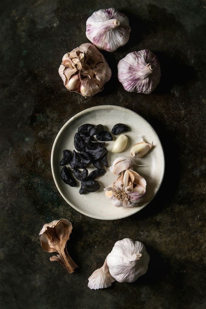 Variety of fresh organic garlic bulbs whole and peeled and cloves of black fermented garlic with ceramic grater over grey spotted background. Flat lay. space. (Photo by: Natasha Breen/REDA&CO/Universal Images Group via Getty Images)