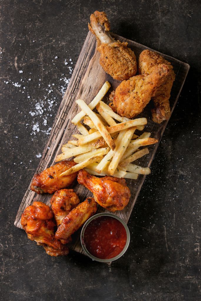 Fast food fried crispy and spicy chicken legs, wings and french fries potatoes with salt and ketchup sauce served on wooden serving board over dark texture background. Top view, space for text. (Photo by: Natasha Breen/REDA&CO/Universal Images Group via Getty Images)