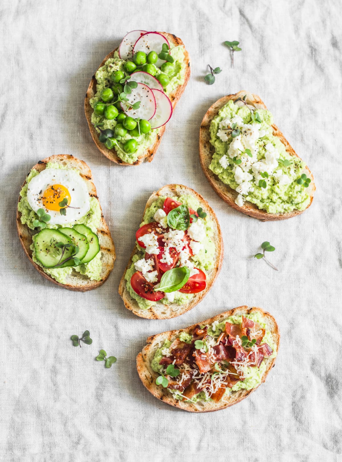 Variation avocado sandwich - with crispy bacon, quail egg, tomatoes, goat cheese, green peas, radish, cucumber. Healthy snack. On a light background, top view