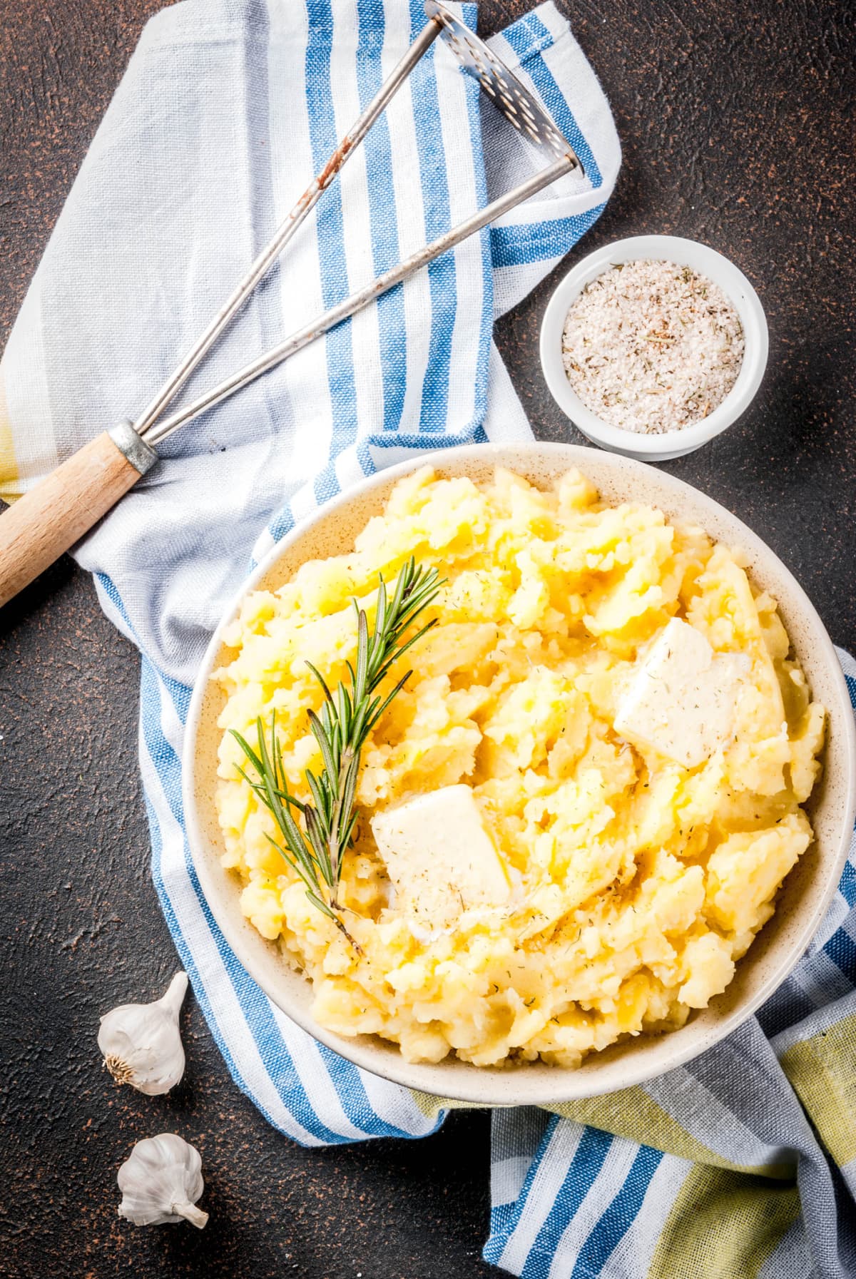 A bowl of mashed potatoes with butter and herbs beside a masher and bowl of seasonings