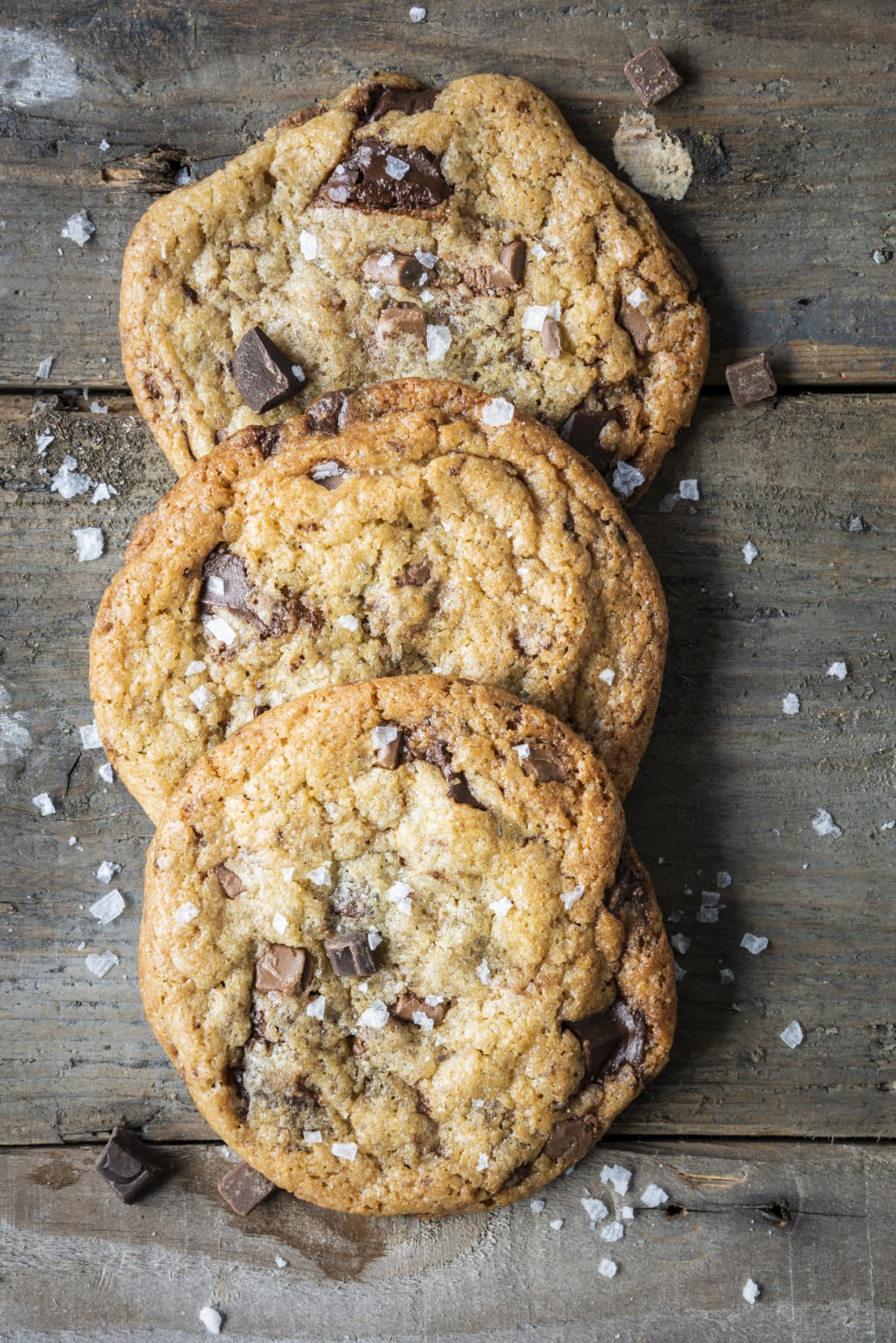 Three freshly baked chocolate chip cookies are lying on a textured wooden background sprinkled with salt. There are loose chocolate chips and salt flakes scattered around the cookies. The chocolate in the cookies has melted leaving the biscuits gooey. Taken from overhead.