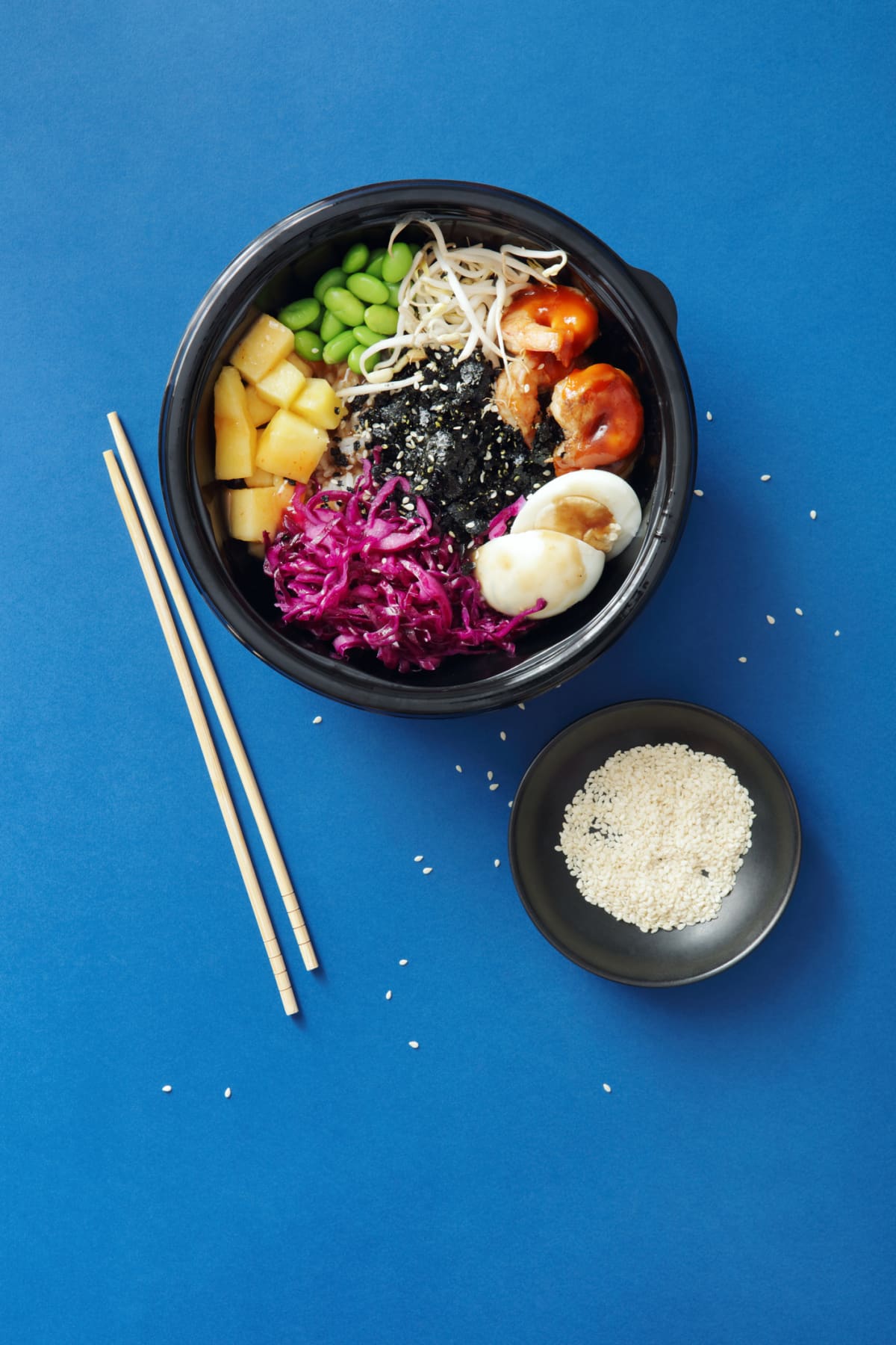 Poke bowls made with grilled prawns, vegetables, cabbage, edamame, bean sprout, wakame, boiled egg, spicy mayo served on a bed of steamed rice. Flat lay top-down composition on blue background. Vertical image with copy space.