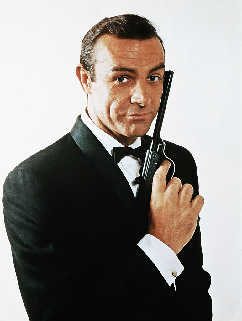 (Original Caption) Waist-up portrait of Sean Connery, as James Bond, caressing the barrel of a gun against the side of his face. Connery is wearing a tuxedo and bow tie and smiling slightly.