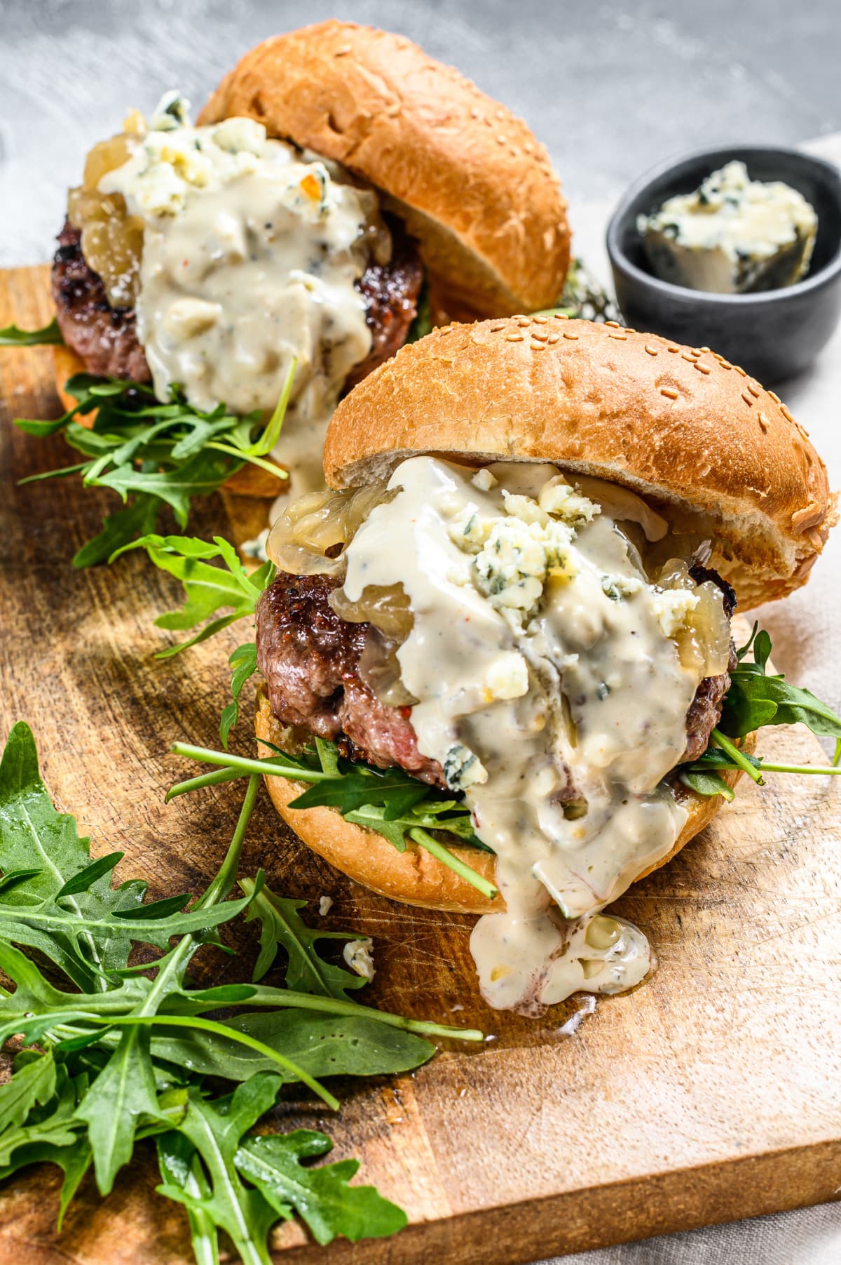 Burgers topped with blue cheese sauce on wooden serving tray with arugula on the side