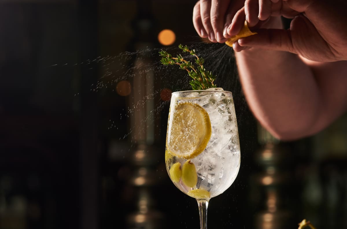 Fingers squeezing lemon peel over spritzer with lemon and herb garnishes 