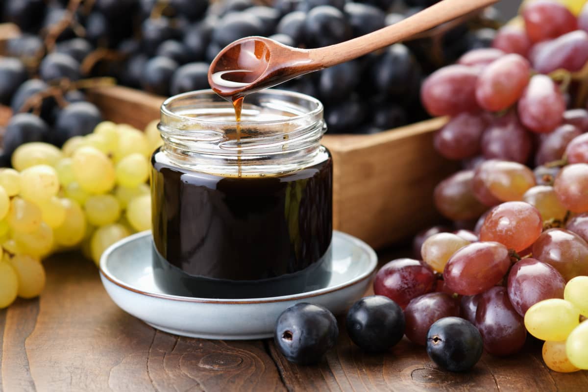 Jar of grape molasses, grape syrup. Sweetener, cough medicine. Black, green and purple grapes on kitchen table.