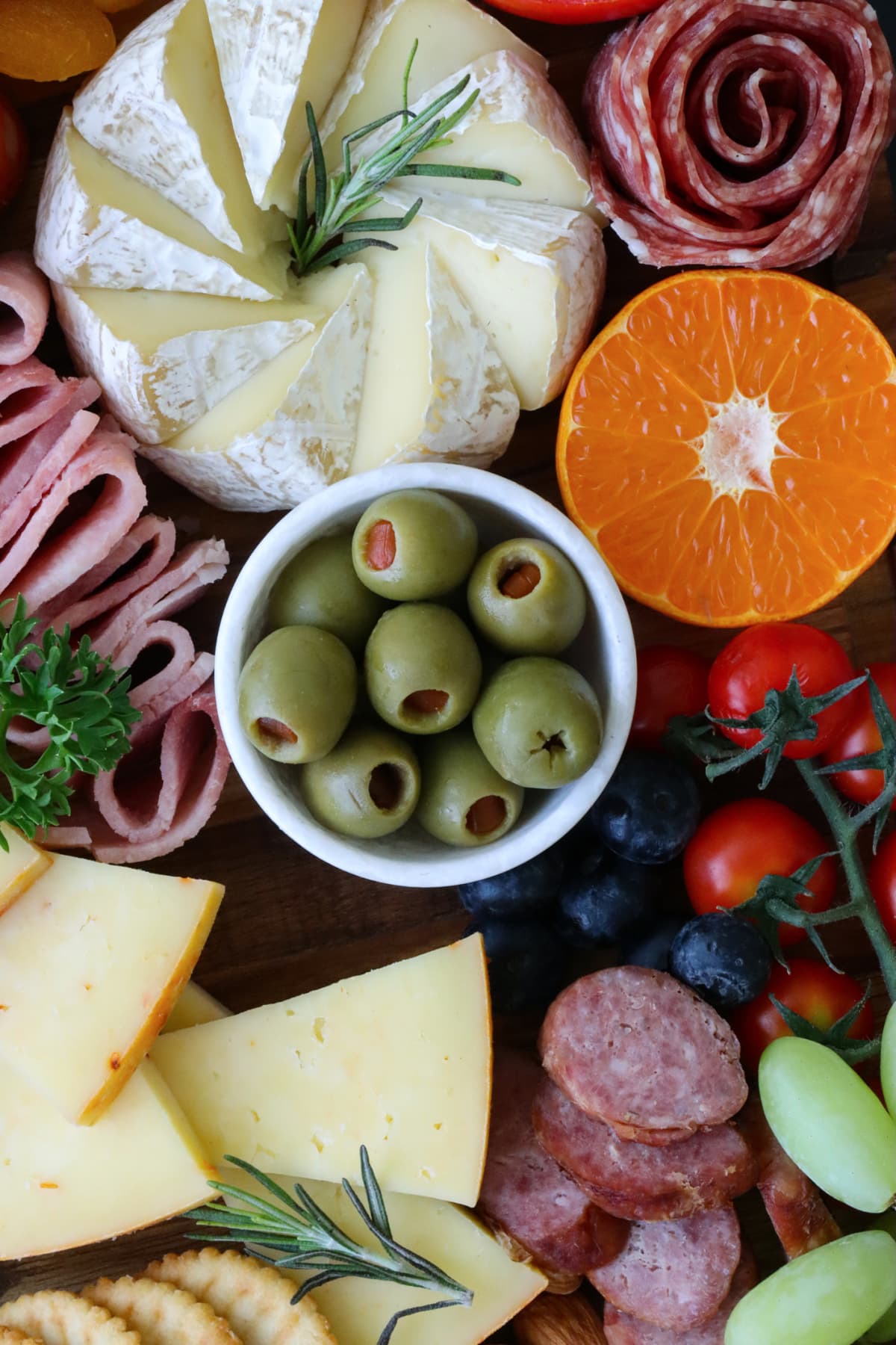 Stock photo showing close-up, elevated view of wooden charcuterie board covered with prepared sliced and chopped ingredients including crackers, ramekin of stuffed olives, salami roses, cherry vine tomatoes, Cheddar cheese, blueberries, orange half, Brie, Parsley and Rosemary herb garnish.