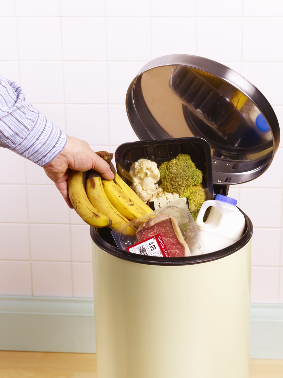 A hand throwing a bunch of bananas into a garbage can overflowing with food