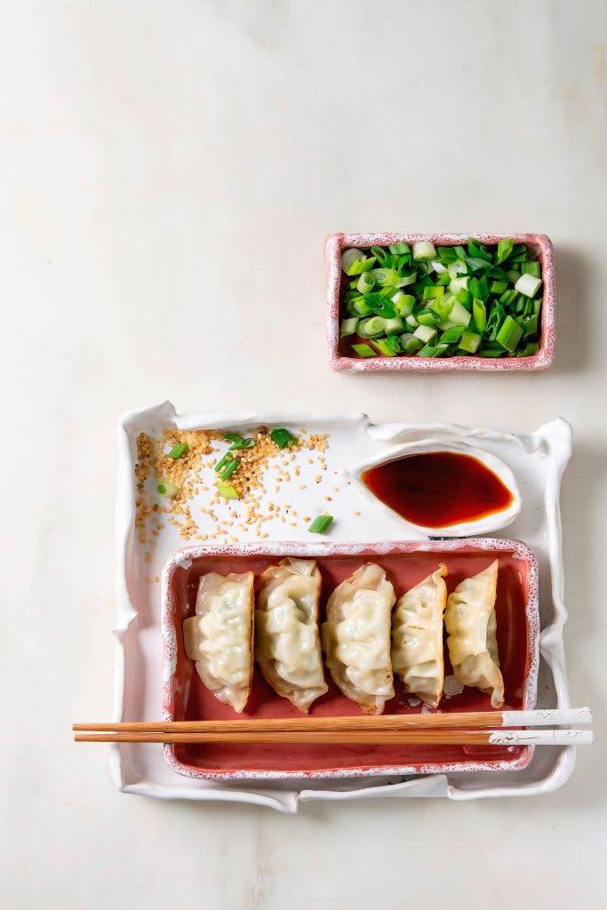 Fried asian dumplings Gyozas potstickers in pink square ceramic plate served with chopsticks, bowl of soy sauce, chopped spring onion over white marble background, Flat lay, space, Asian dinner. (Photo by: Natasha Breen/REDA&CO/Universal Images Group via Getty Images)