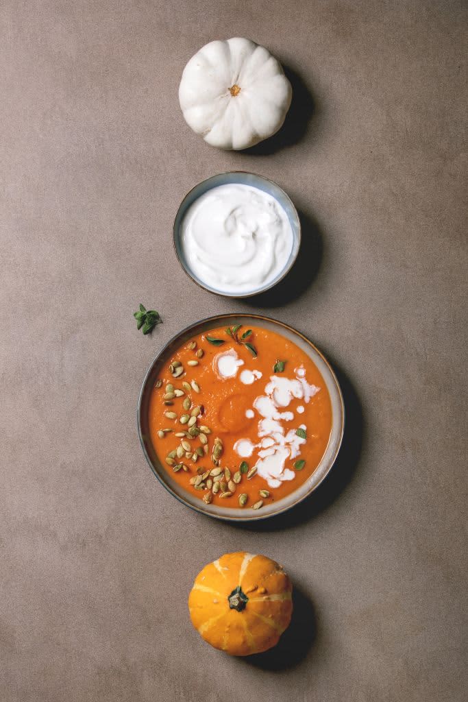 Pumpkin soup with cream and seeds in ceramic bowl, served with sour cream, uncooked pumpkins in row over brown texture background. Flat lay, space. (Photo by: Natasha Breen/REDA&CO/Universal Images Group via Getty Images)