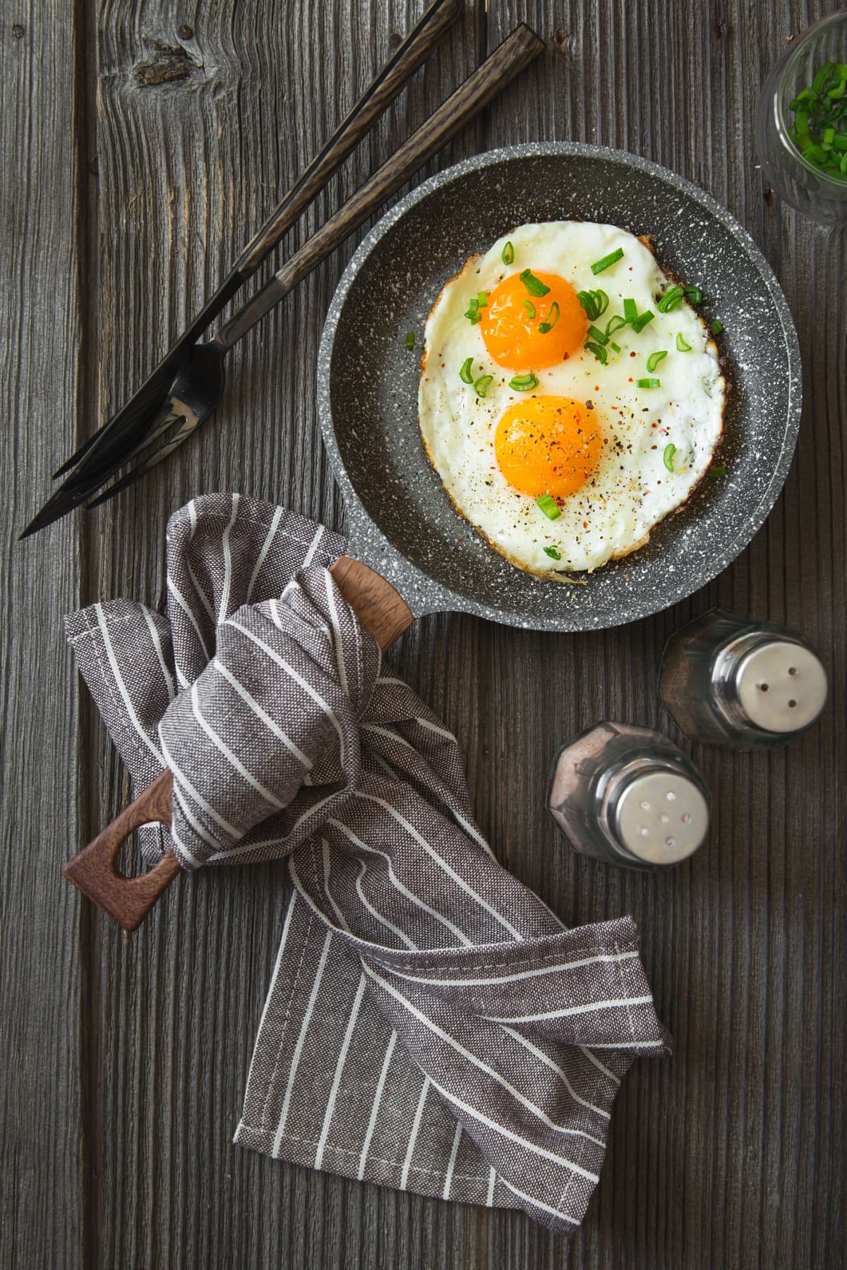Fried eggs with green chives chopped for breakfast. Two fried eggs in a small pan on the dark wooden background, top view. Flat lay. Rustic style.