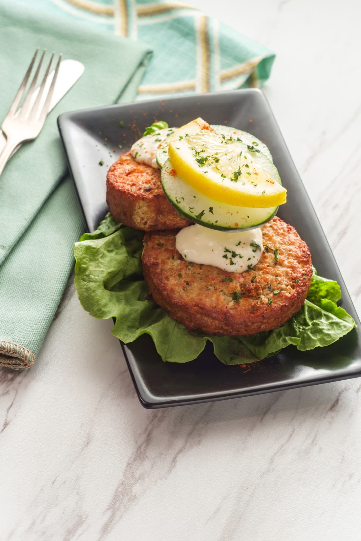 Two crab cakes on a plate topped with sauce, cucumber, and lemon