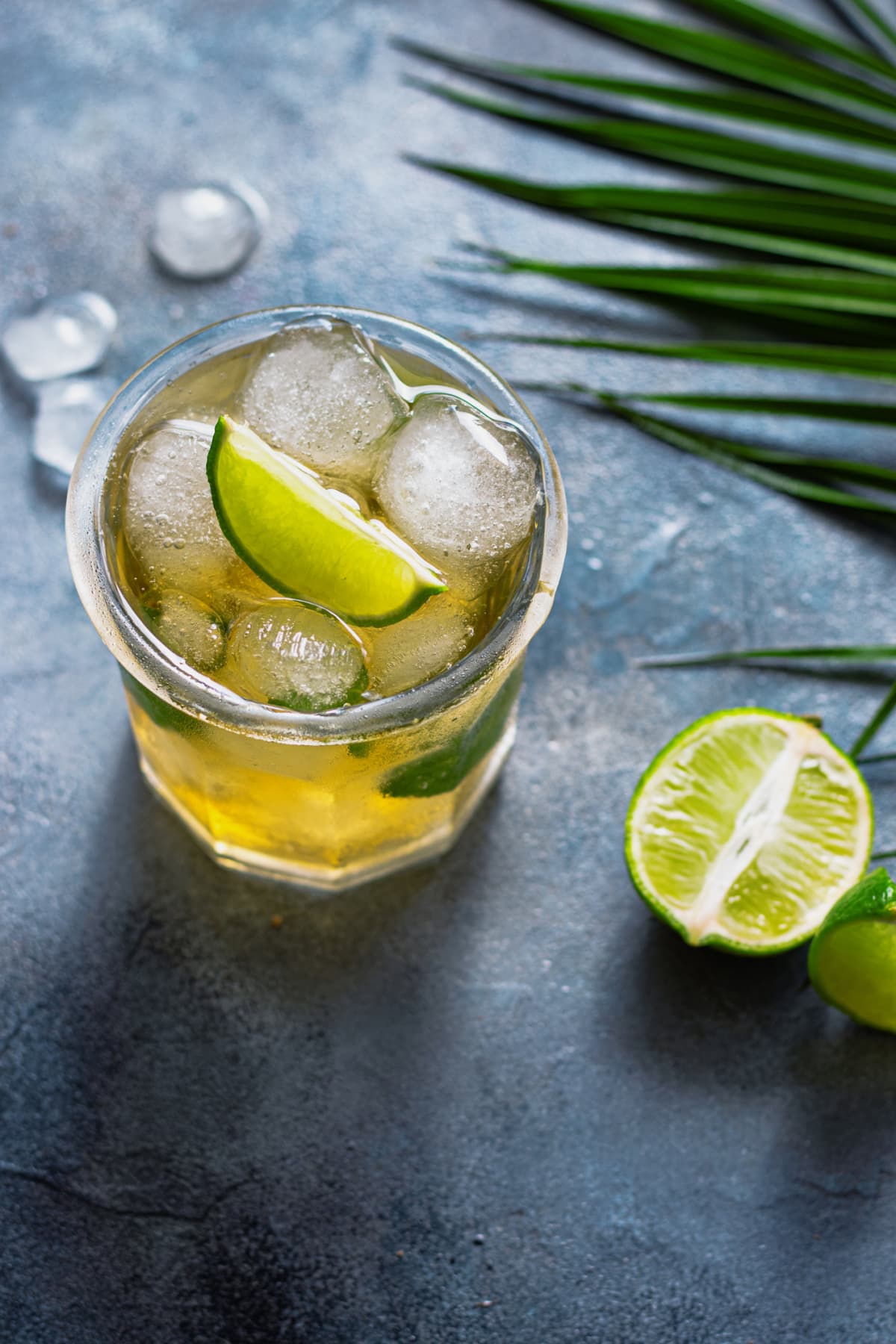 Green ice tea with ice cubes, lime, and mint leaves in the glass with palm leaves on gray stone background