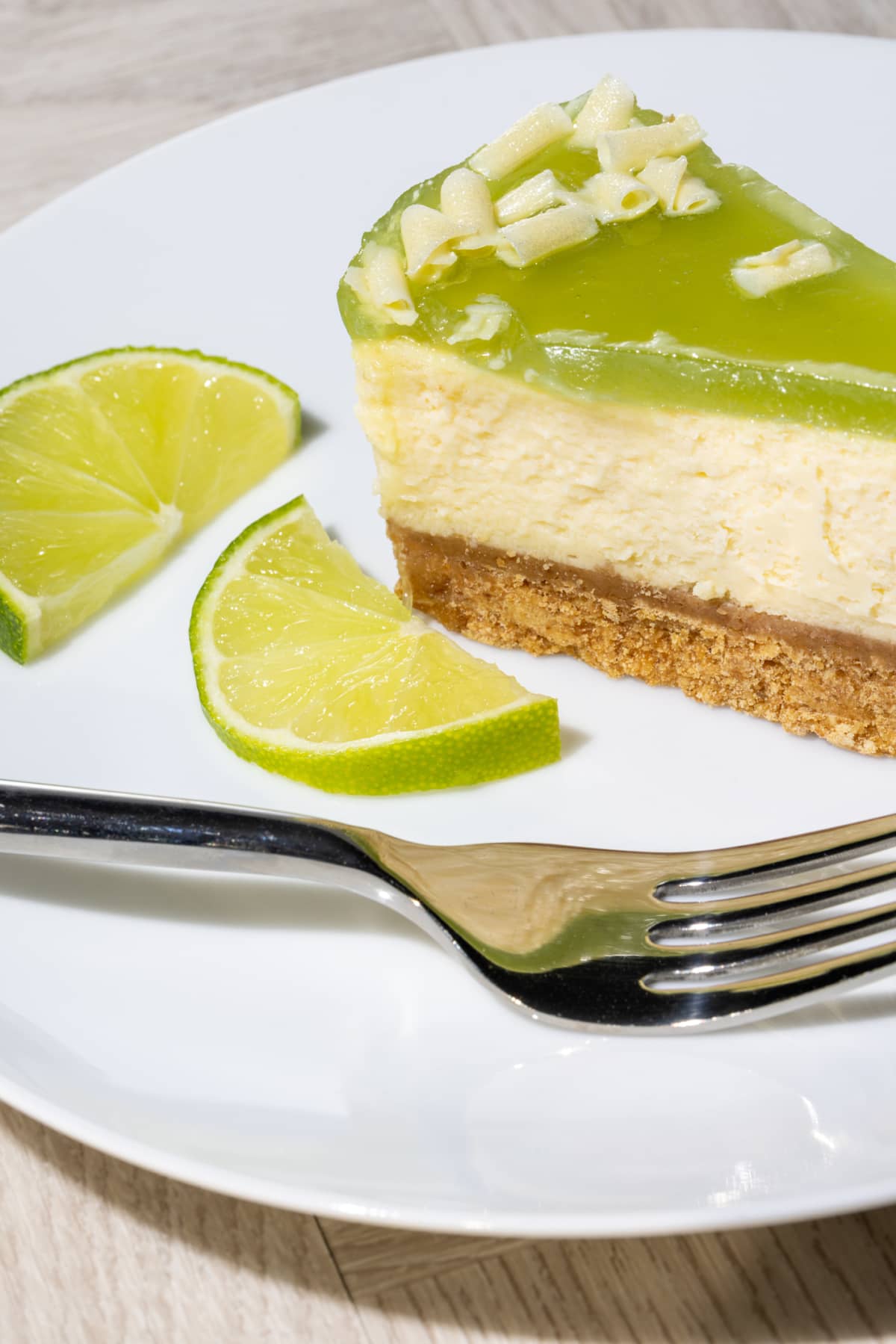 Lime cheesecake slice. Close-up of a delicious sweet citrus dessert. One serving of a baked key lime cheesecake with digestive biscuit base served on a white plate with fork and green lime slices