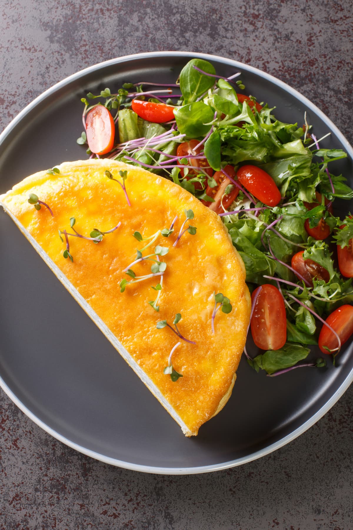 Lunch or dinner egg dishes is the famous Poulard Omelette with vegetables salad closeup on the plate on the dark background. Vertical top view from above