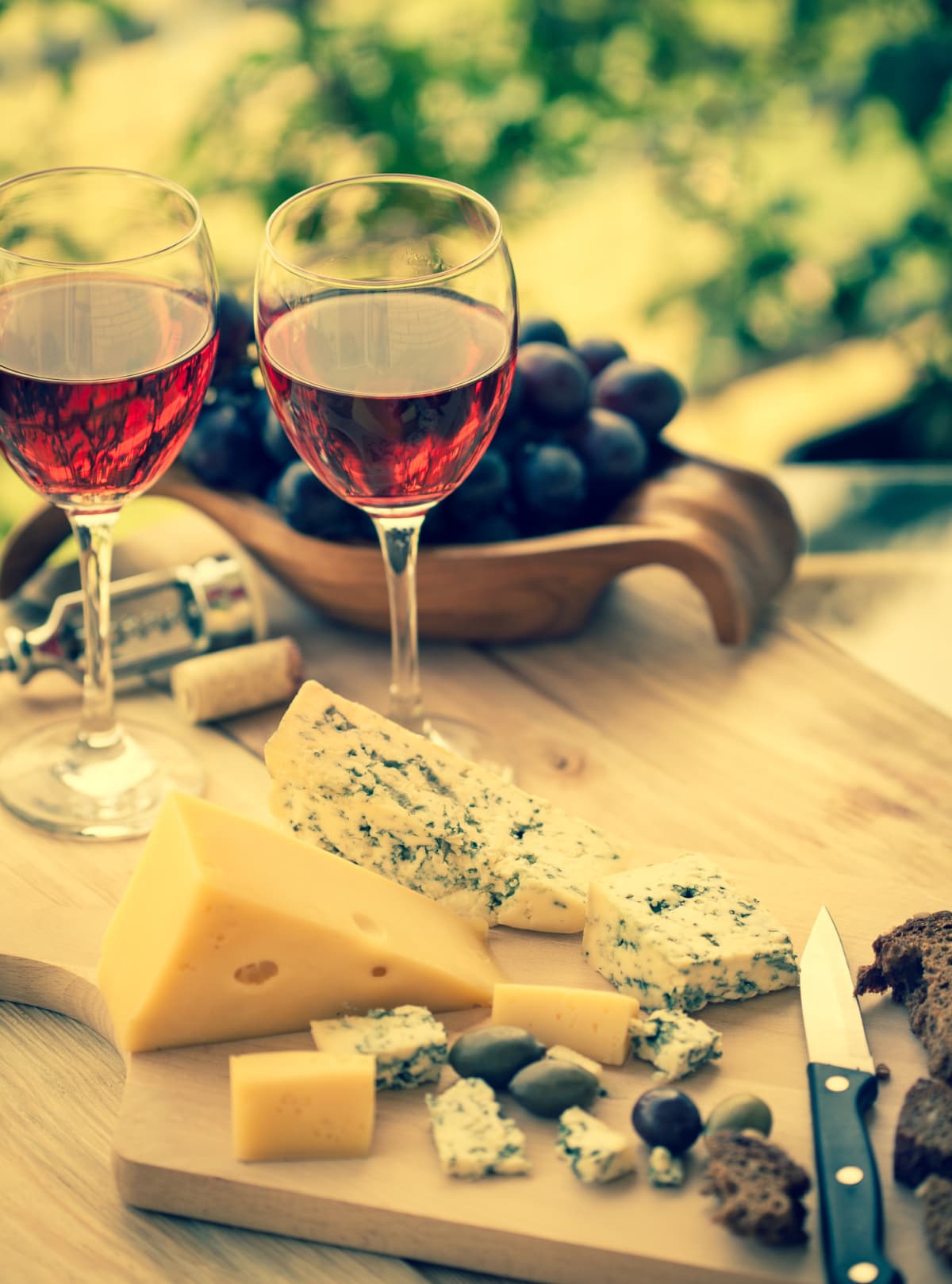 two glasses of red wine with grapes and cheese on a wooden cutting board