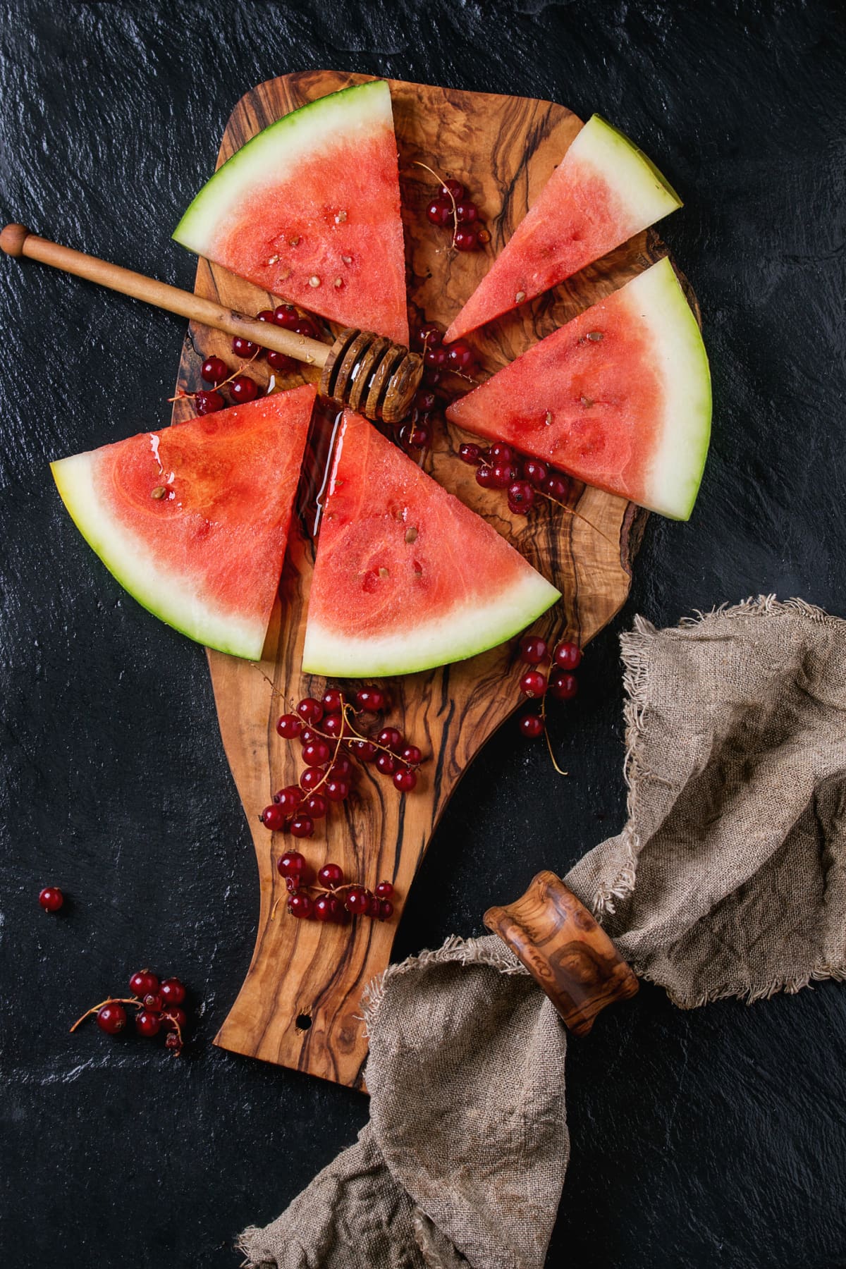 Sliced watermelon with red currant and honey, served on olive wood chopping board with honey dipper and sackcloth napkin over black slate background. Top view. Healthy dessert concept. 