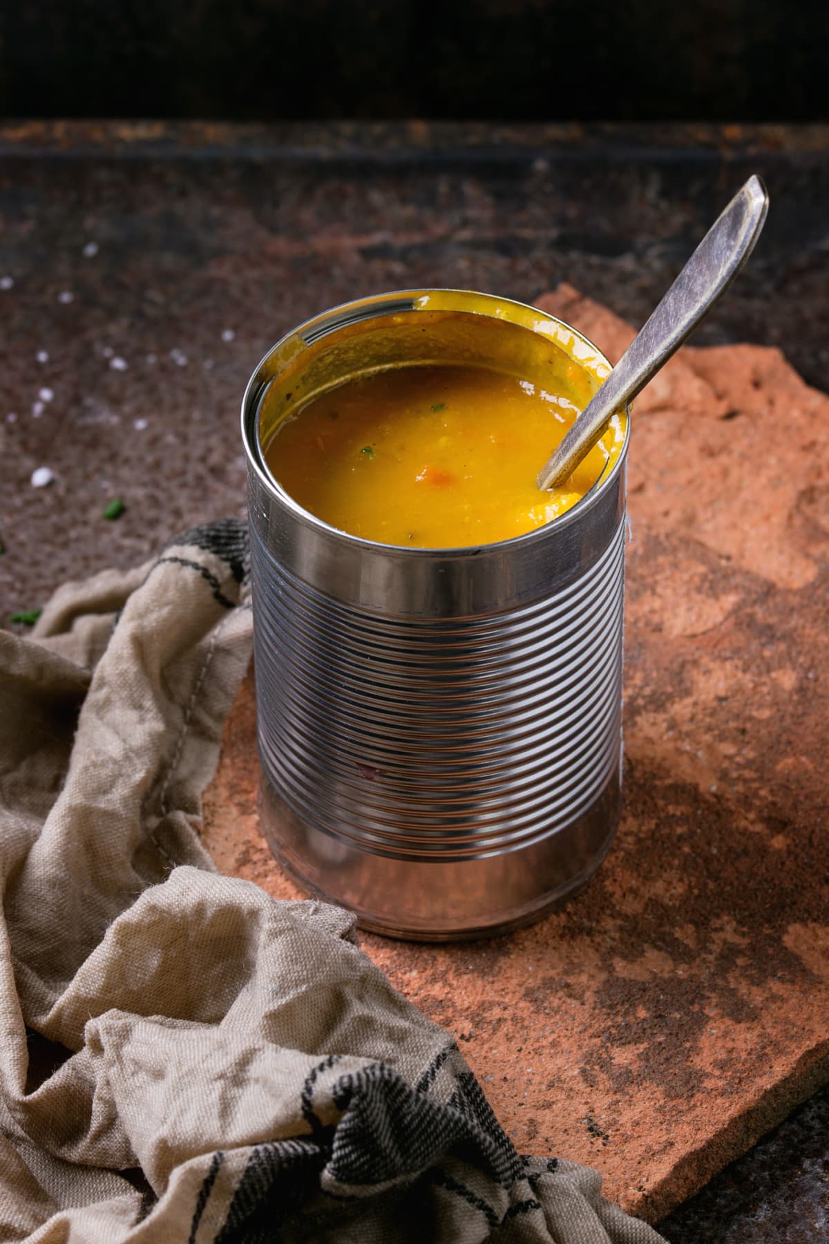 Tin can of carrot soup with spoon, standing on kitchen towel  and clay board over old rusty iron background. Dark rustic style