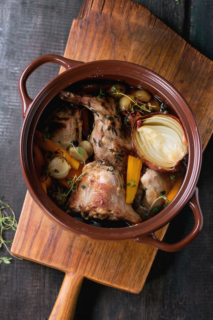 Brown ceramic pot with stewed rabbit with bouillon, vegetables and herbs, served on wooden chopping board over old wood table. Rustic style. Flat lay. (Photo by: Natasha Breen/REDA&CO/Universal Images Group via Getty Images)