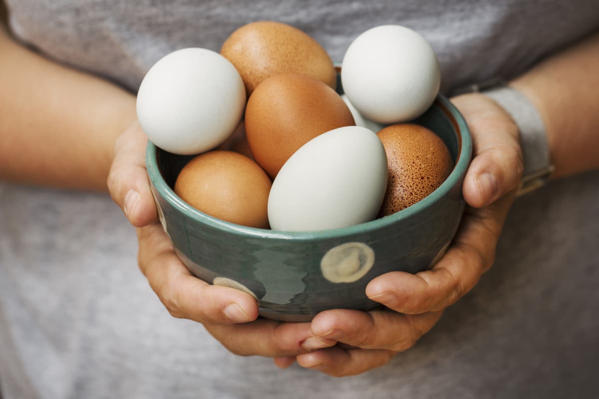 A detail of cracked egg falling into the pan as woman holds egg shells in both hands.
