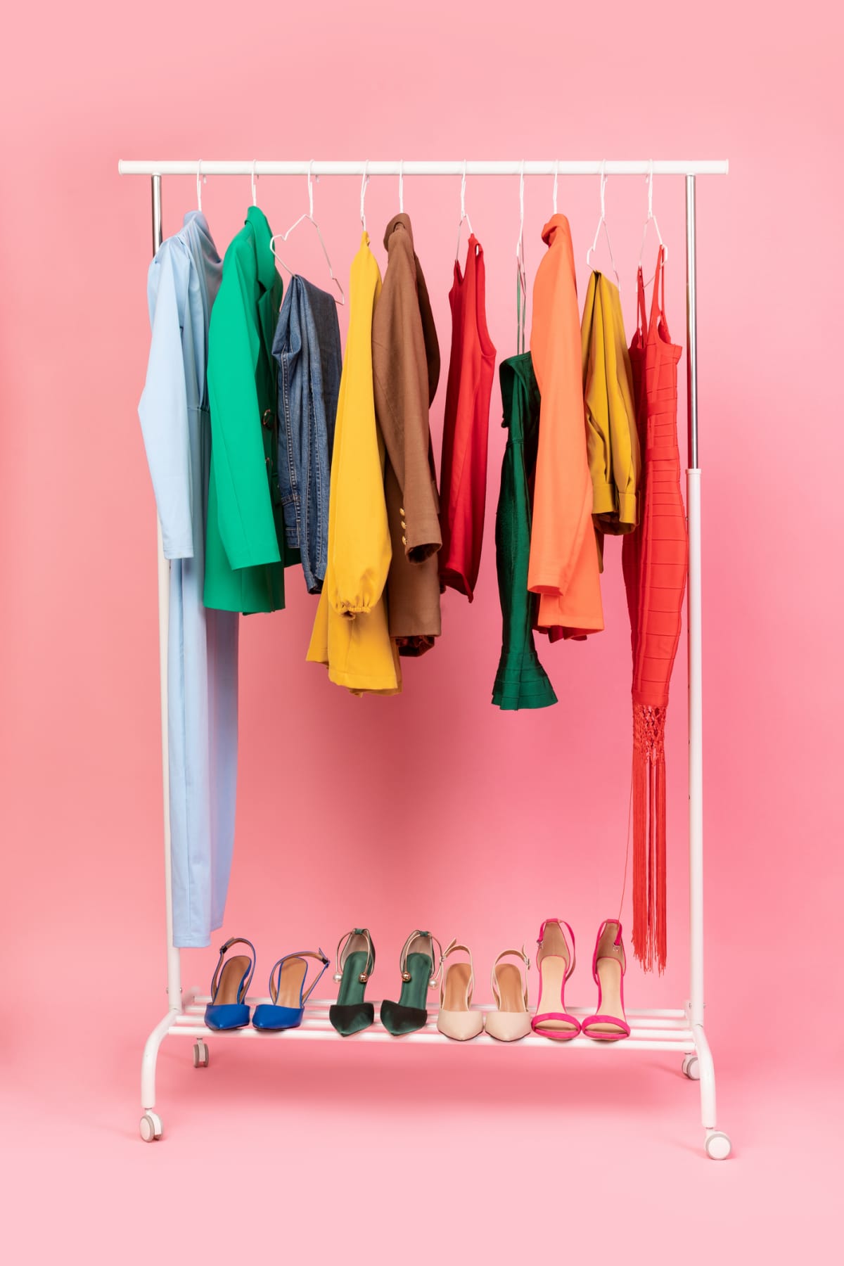  Vertical Shot Of Clothing Rail With Bright Trendy Clothes Hanging On Hangers And Shoes Over Pink Studio Background In Empty Room. 