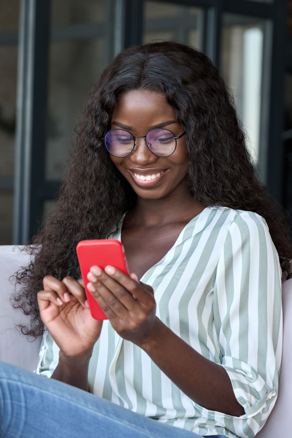 Young happy black African woman wearing glasses using mobile digital apps on cell phone tech device online shopping or dating sitting on couch at home, texting on smartphone, writing social post.