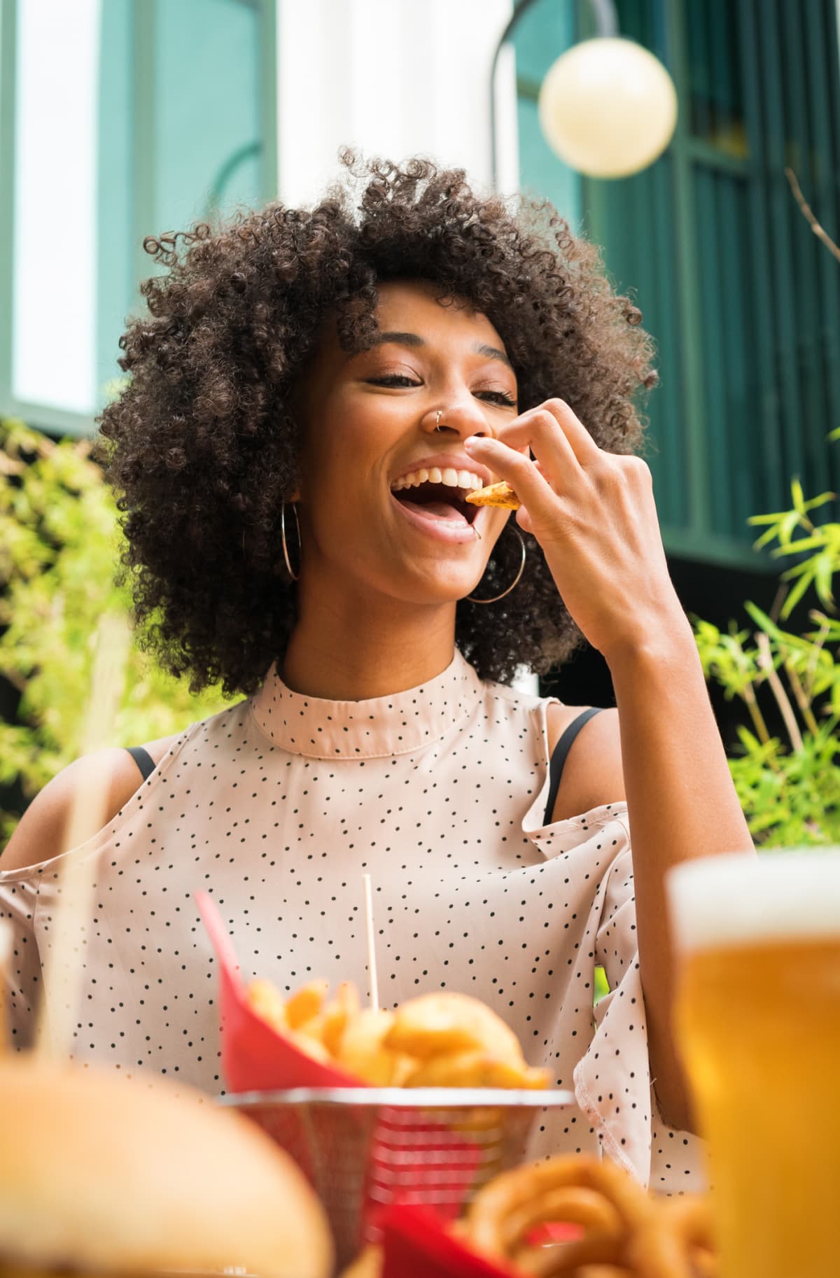 Smiling beautiful young happy Black woman with nose piercing eating potato wedges in a pub in a low angle view over food and glasses of beer