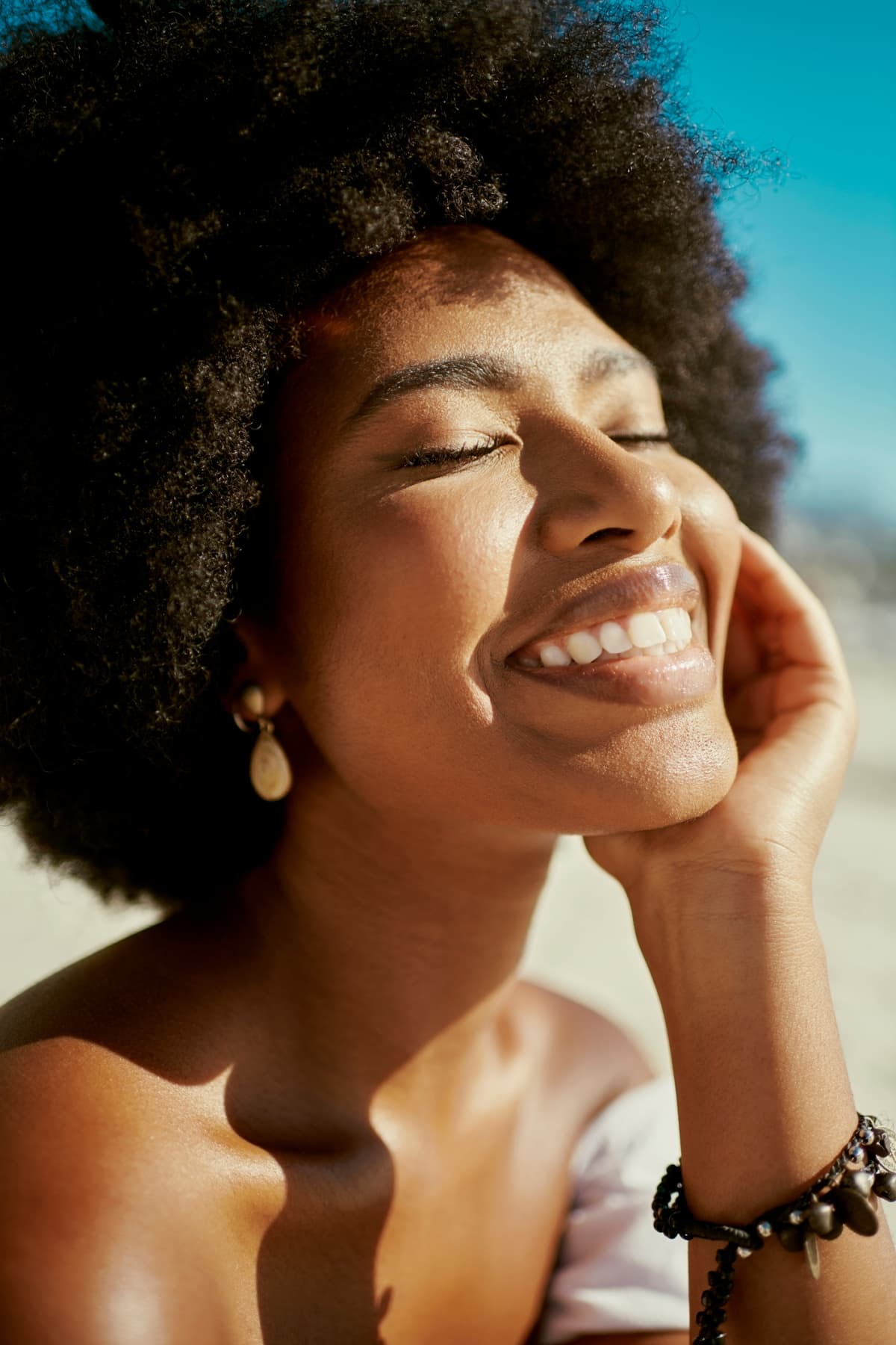 Happy, smiling and beautiful woman with an afro closed eyes closeup. Gorgeous, attractive and desirable young black female wearing makeup with good skin looking stunning in the sun on hot Spring day
