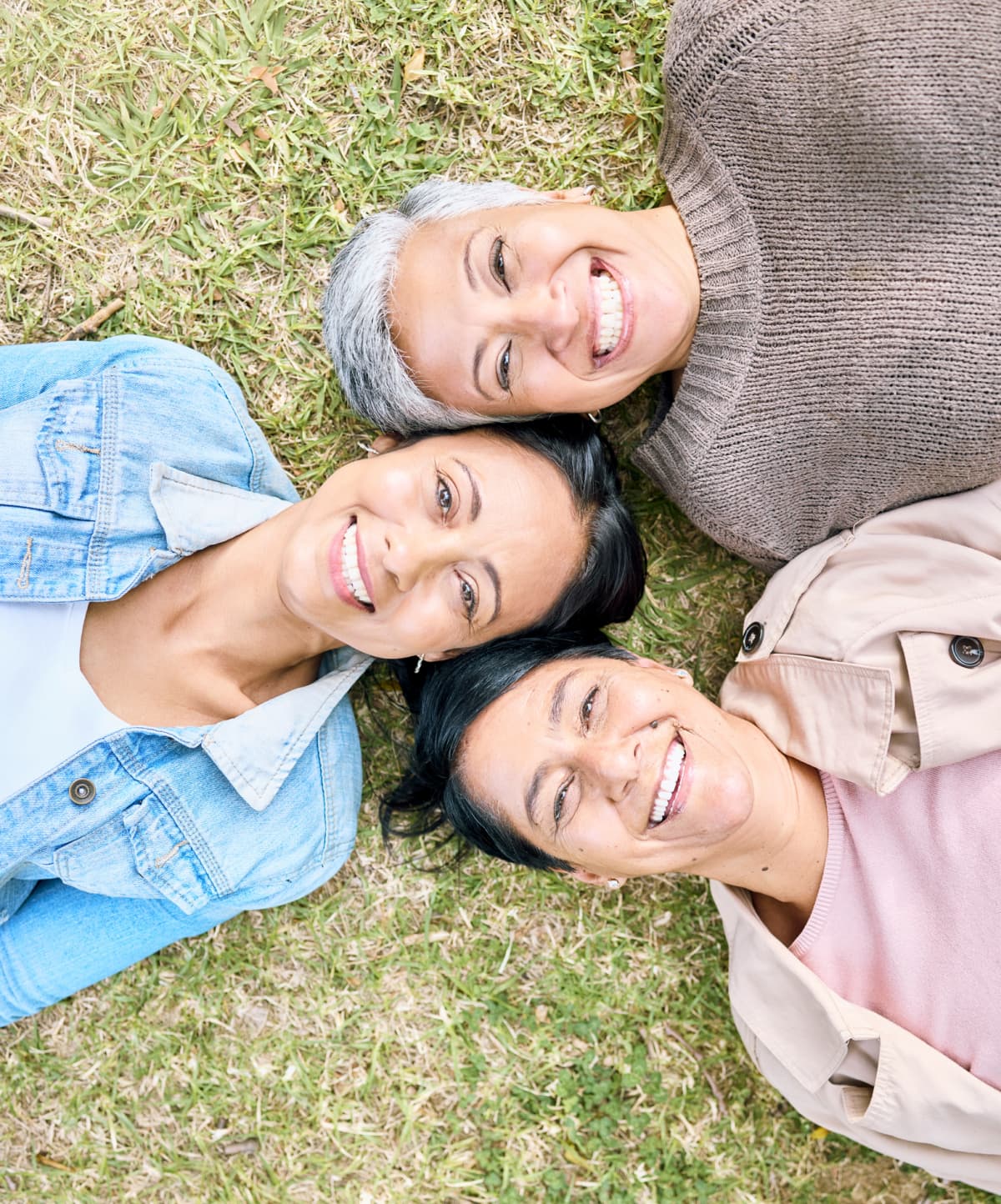 mother, grandmother, and adult daughter relax on the grass, laughing