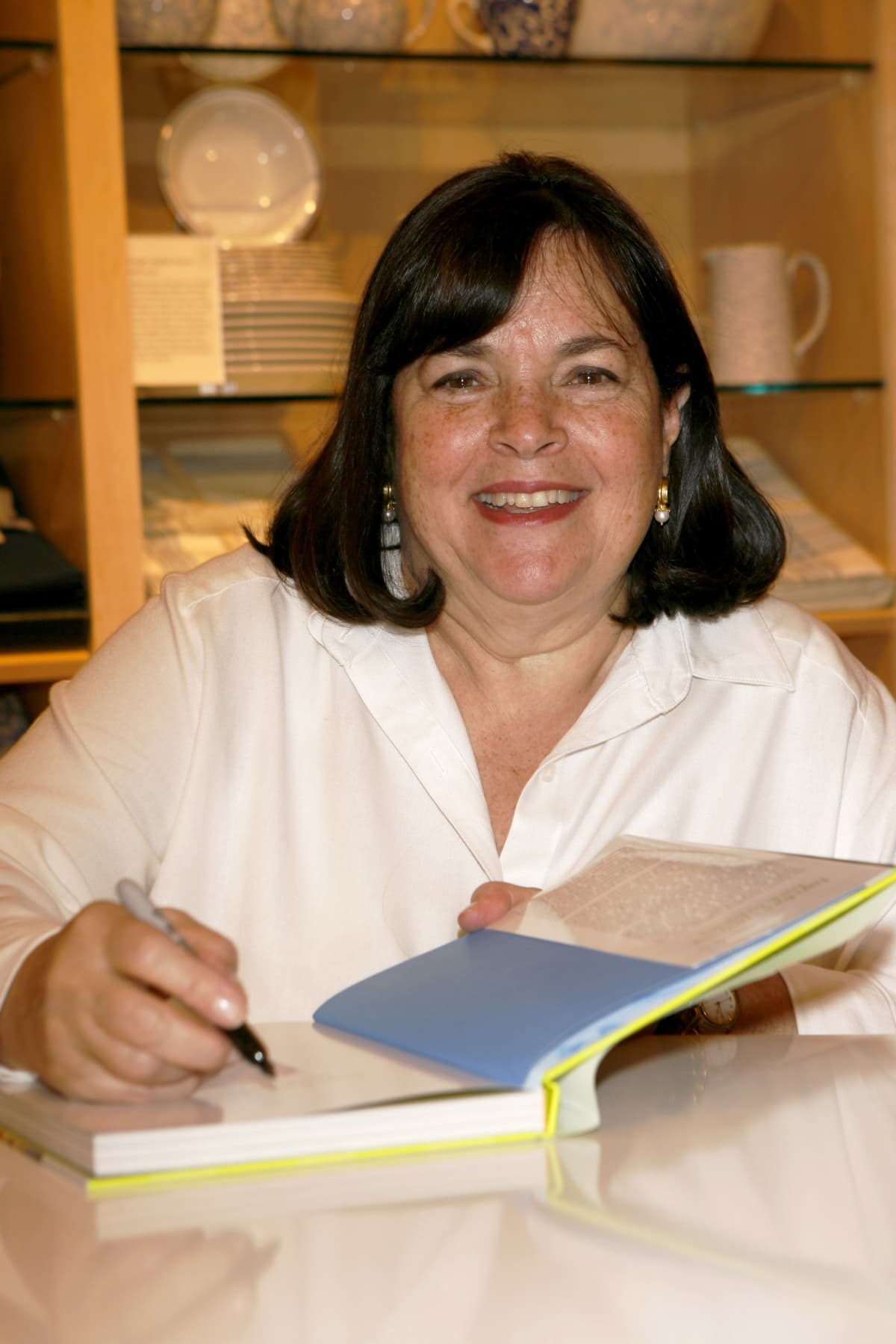 Ina Garten smiling and signing a book with her autograph