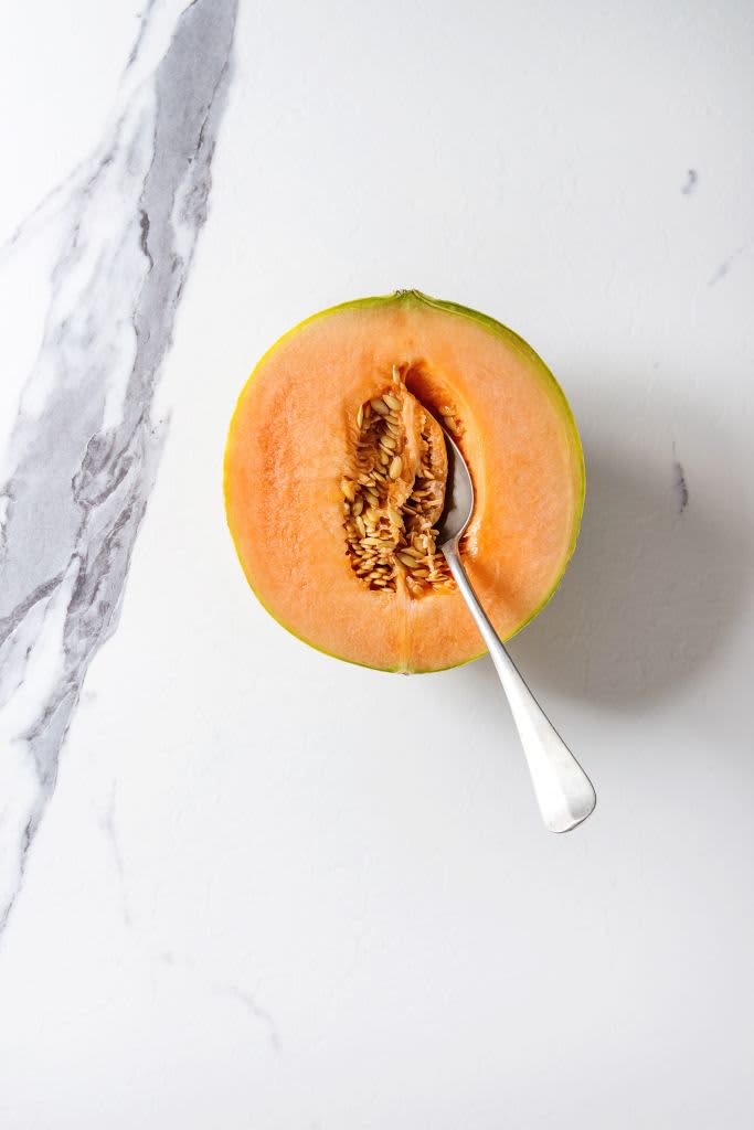 Half of ripe organic Cantaloupe melon with seeds and spoon inside over white marble texture background