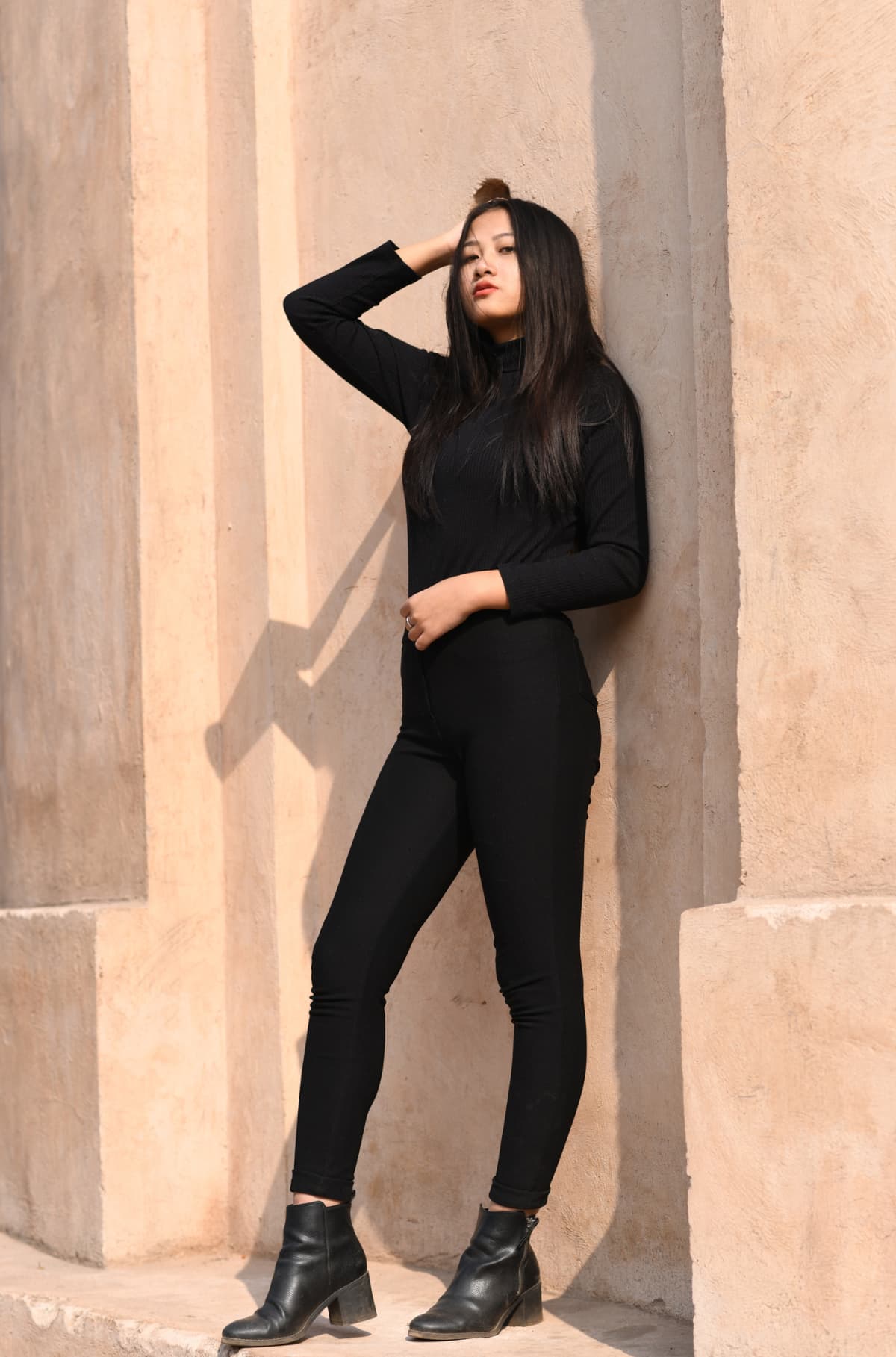 Woman poses against a wall wearing a black bodysuit with black leggings and boots