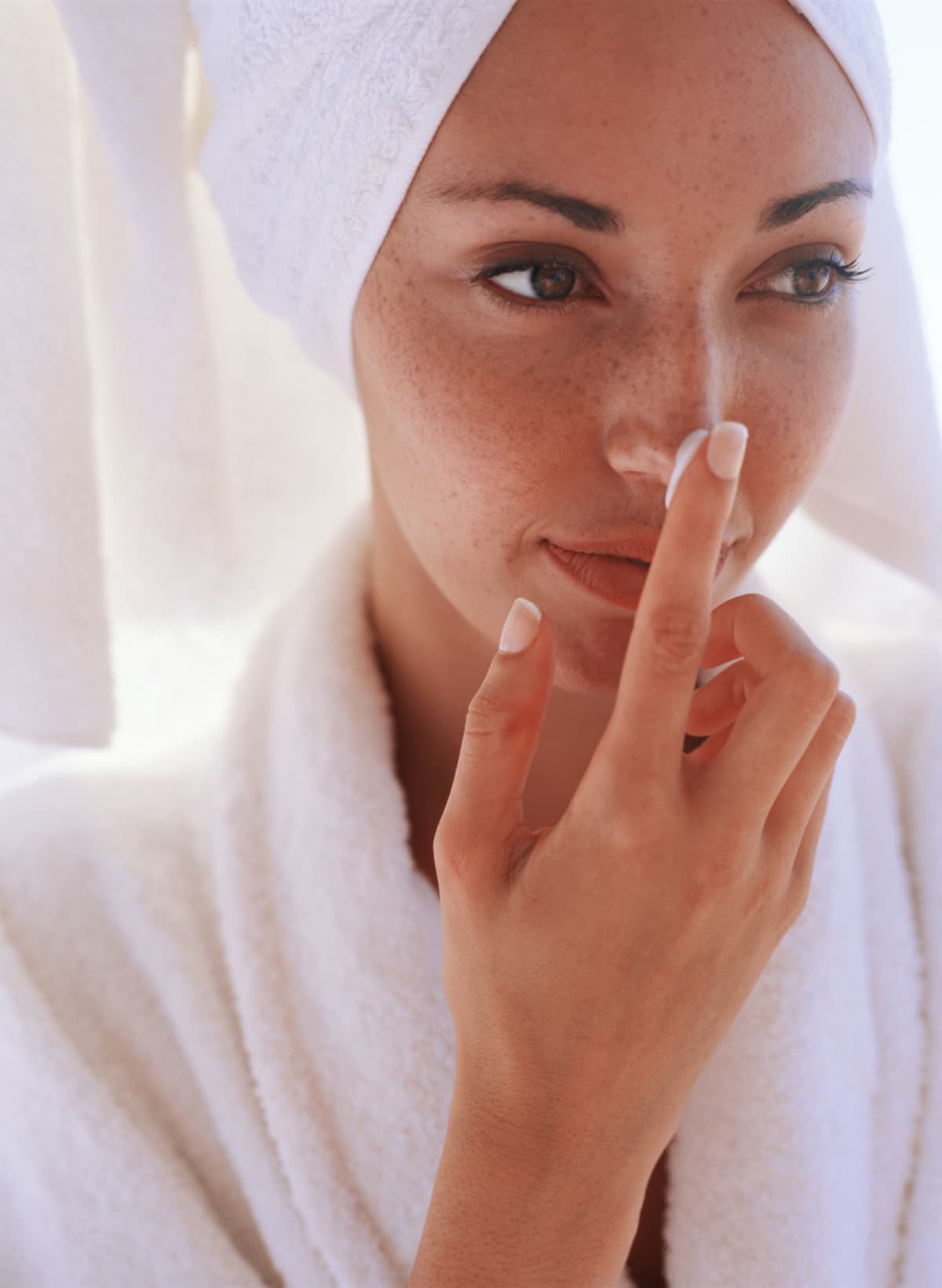 Woman wearing a bathrobe and bathtowel wrapped around her head applies moisturizer to her nose
