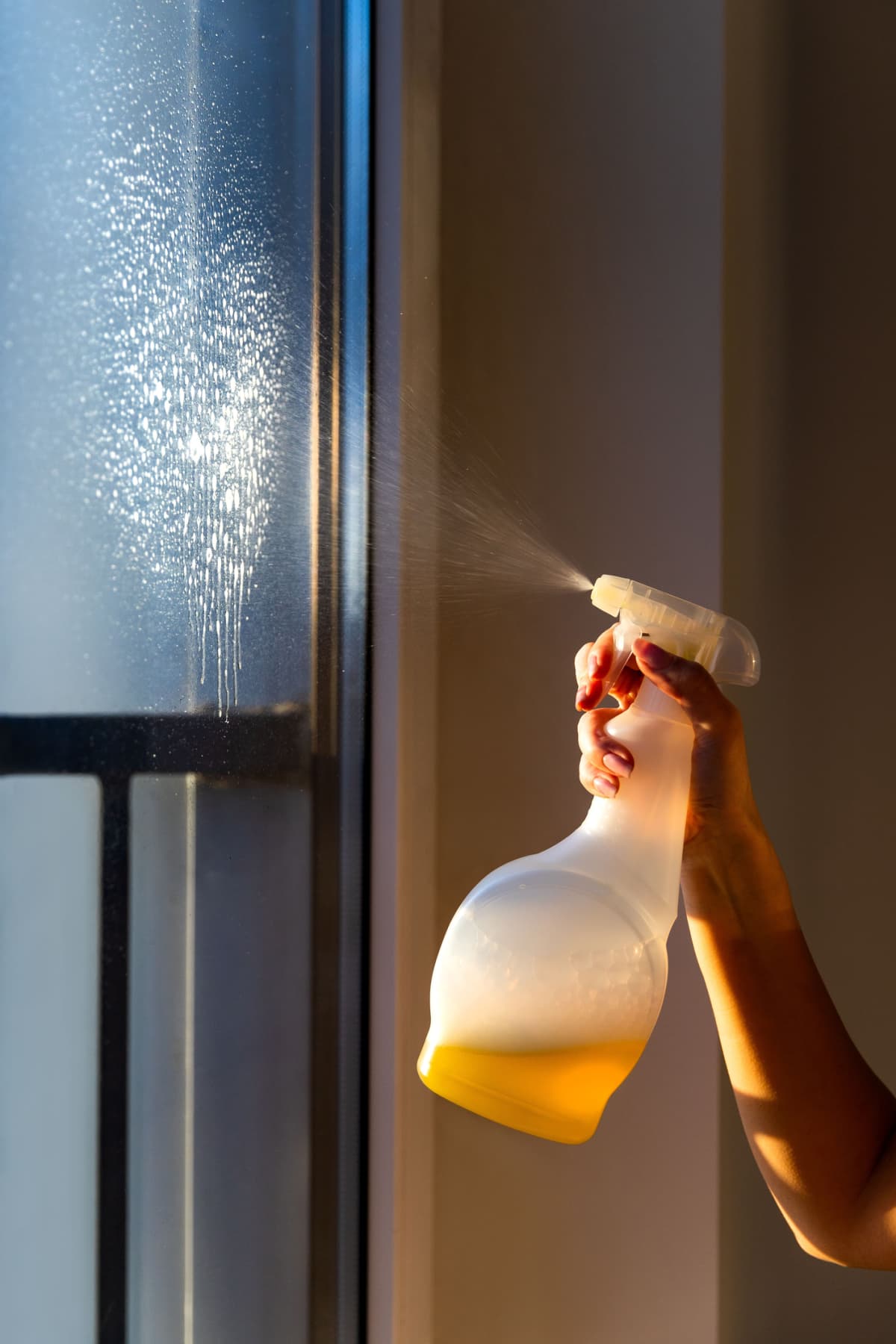 A woman cleaning a window using cleaning sprayer with yellow washing liquid.