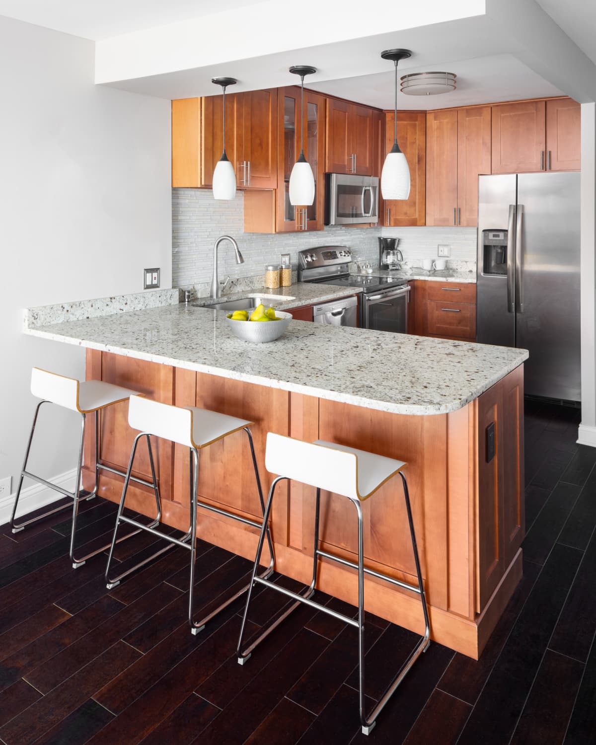 An apartment kitchen with wood cabinets, stainless steel appliances, and pendant lights hanging above the granite countertops.