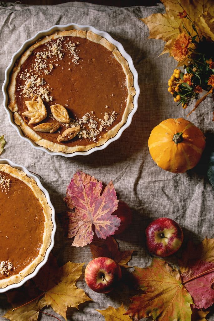 Two pumpkin pies on table with tablecloth, leaves, and pumpkin