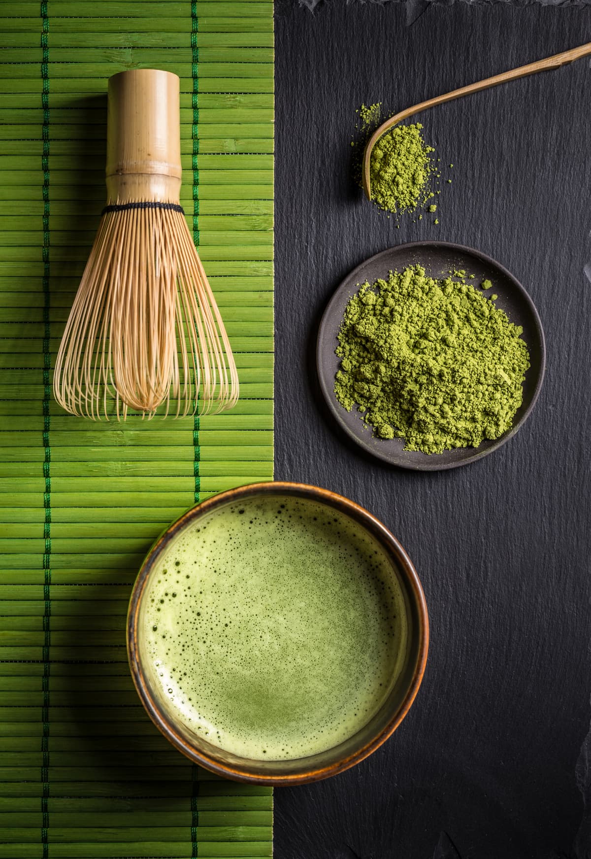 Japanese matcha green tea powder in craft ceramic bowl, couple of cups, bamboo whisk over dark texture background. Flat lay, space. (Photo by: Natasha Breen/REDA&CO/Universal Images Group via Getty Images)