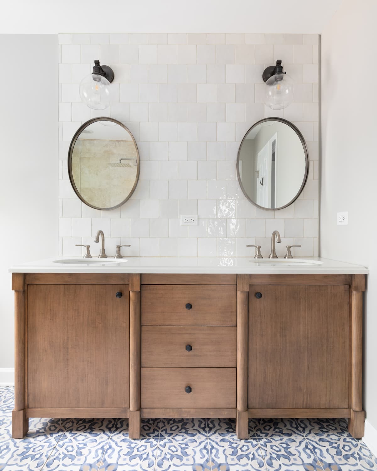 A cozy bathroom with a patterned tile floor, natural wood vanity, tiled backsplash, and lights mounted above circular mirrors