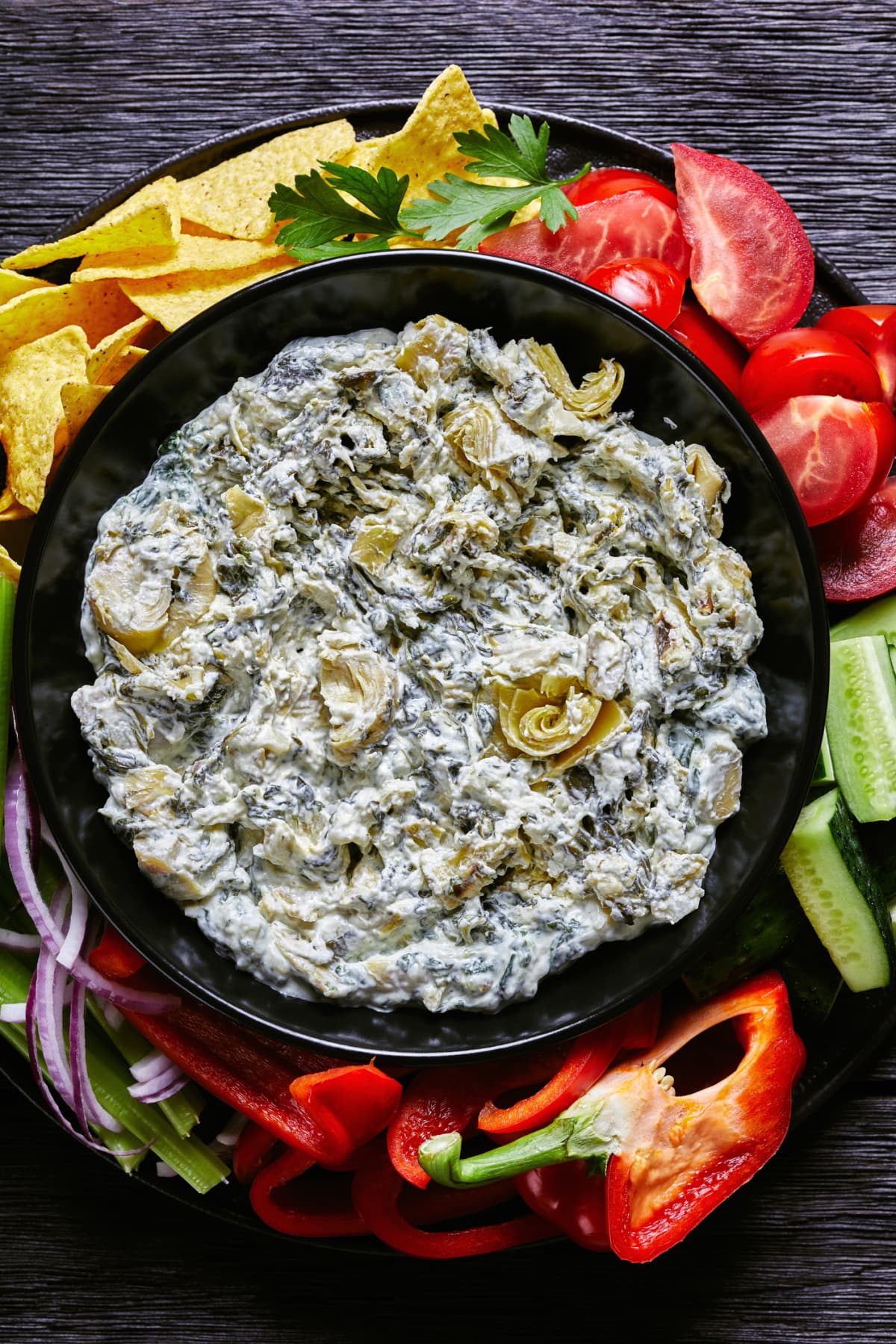 Spinach Artichoke leafy cheese dip in a bowl with tortilla chips, cucumber, celery sticks, tomato slices and red bell pepper.