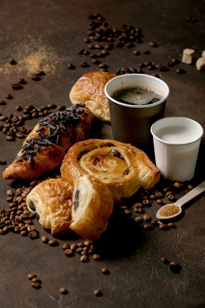 Variety of traditional french puff pastry raisin and chocolate buns. croissant with cups of coffee americano and milk. coffee beans. recycled wooden spoon of sugar over dark texture background. (Photo by: Natasha Breen/REDA&CO/Universal Images Group via Getty Images)
