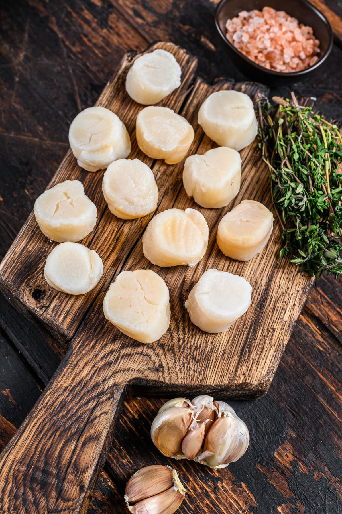 Raw scallops on wooden cutting board with garlic, herbs, and salt on the side