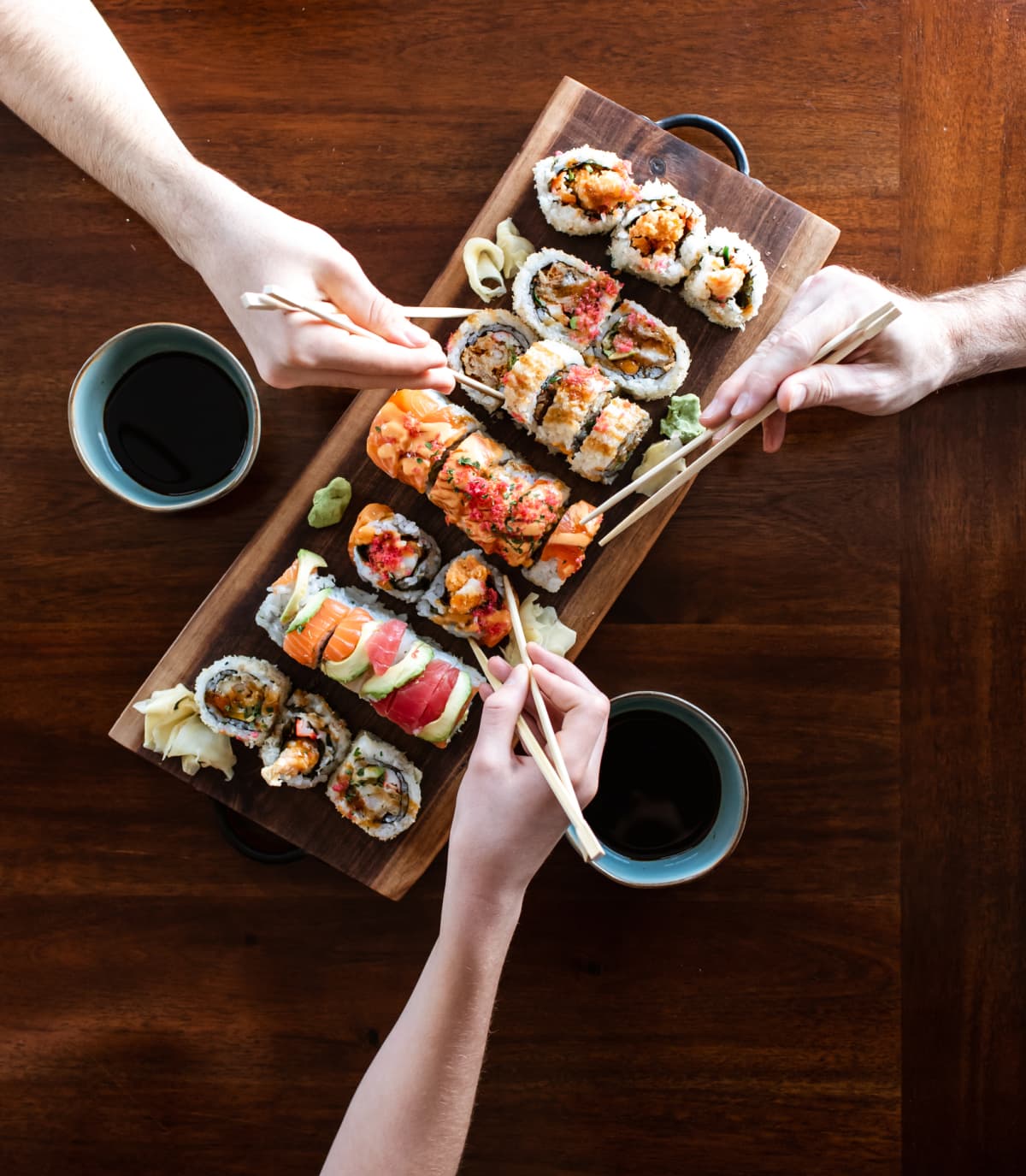 Hands using chopsticks to pick up pieces of sushi rolls from a wooden platter