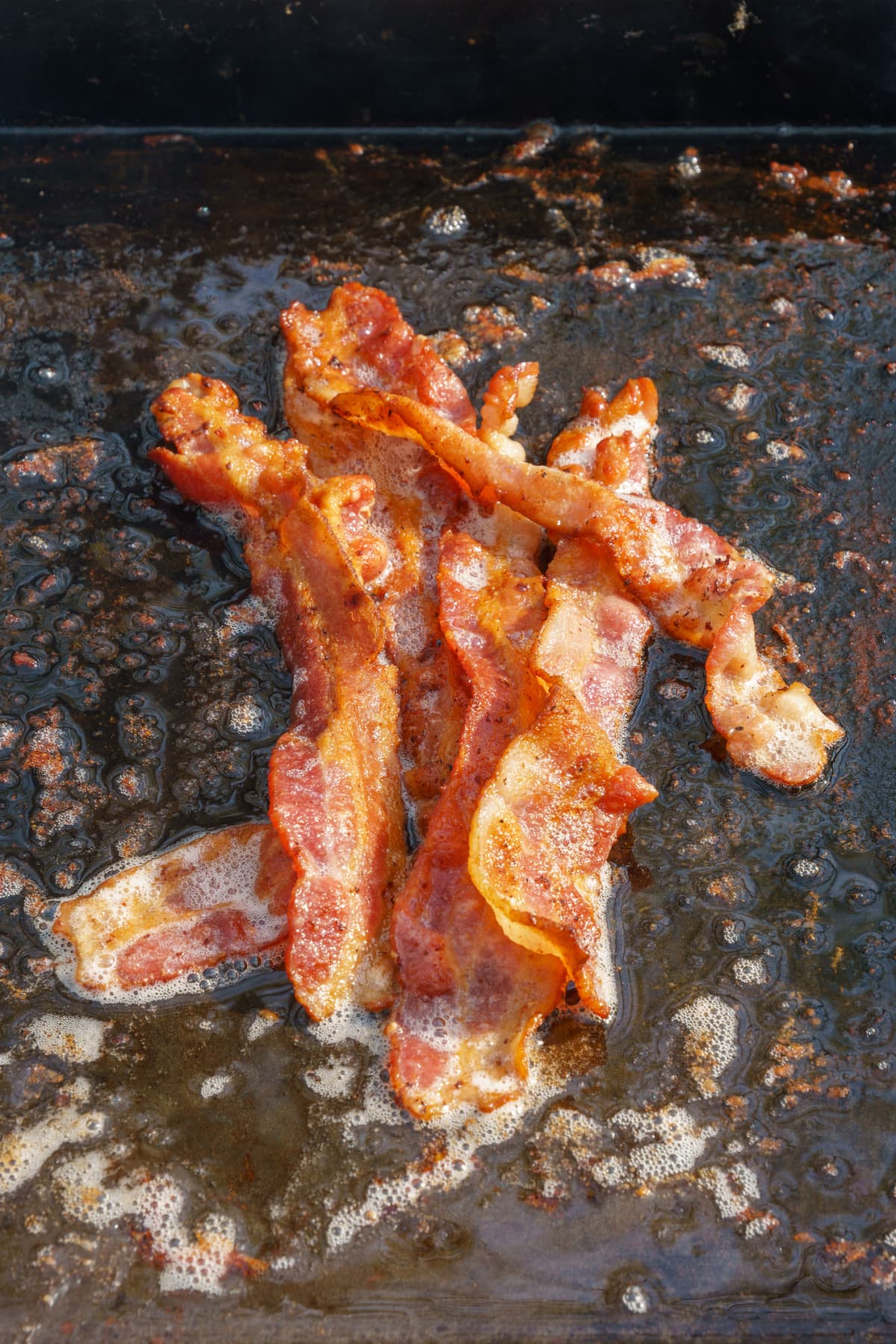 Bacon frying in grease on an outdoor camping grill. The pieces are piled together in the center.