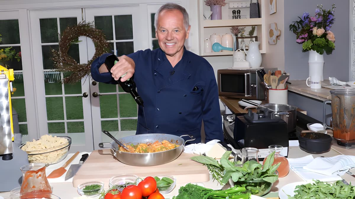 Wolfgang Puck smiling as he adds oil to a pan of cooking food