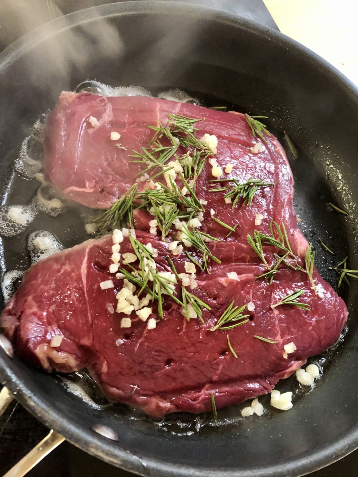 Stock photo showing an elevated view of a smoking hot frying pan containing a raw rump steak seasoned with Garlic and Rosemary.