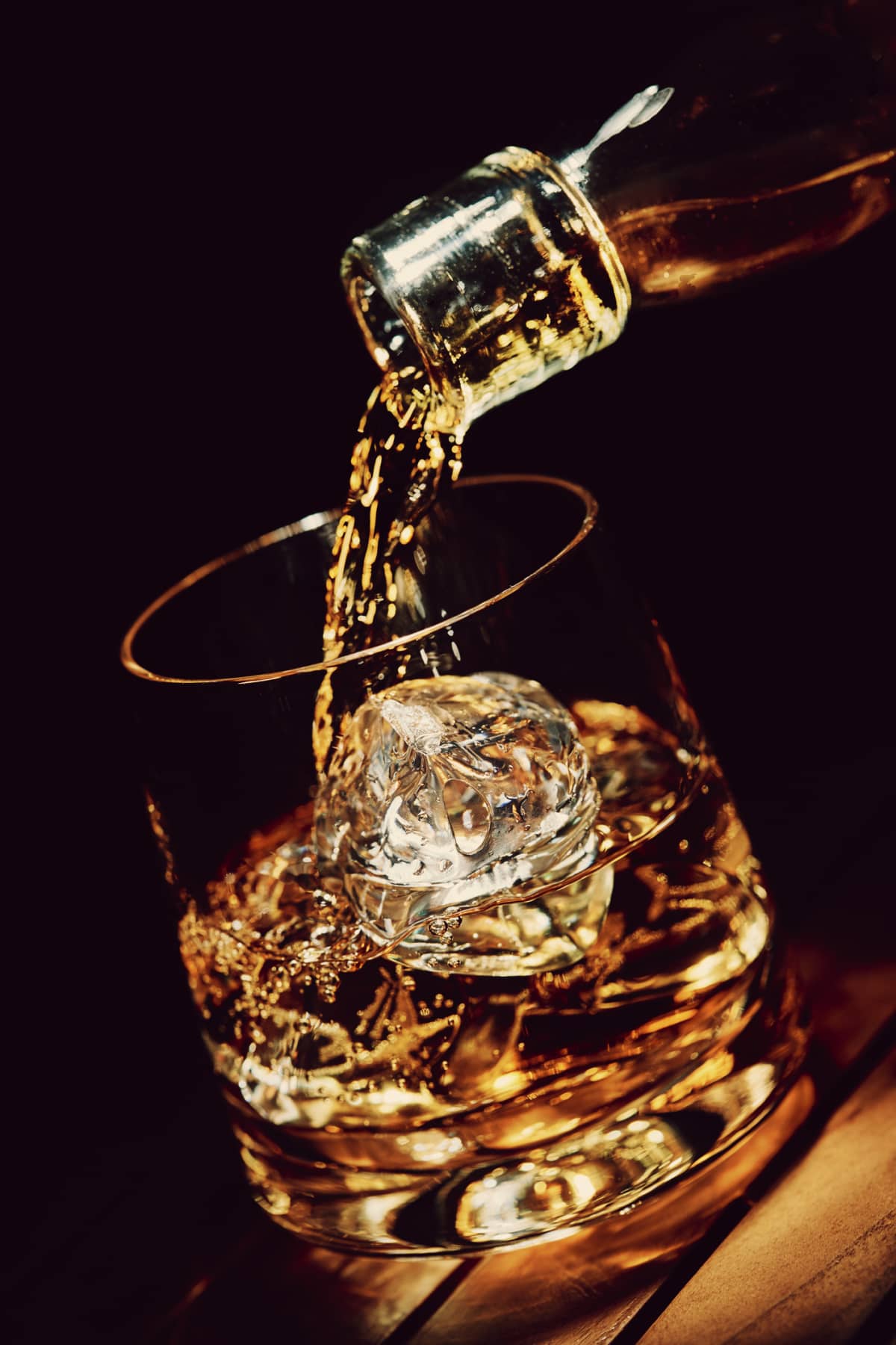 This is a close up photo of whiskey or bourbon splashing out of a shot glass surrounded by ice cubes on a black reflective surface Taken in the studio. This image was photographed at 1/8000th of a second completely freezing the splash.  There is a warm golden light giving it a classy feel with a glow in the background.