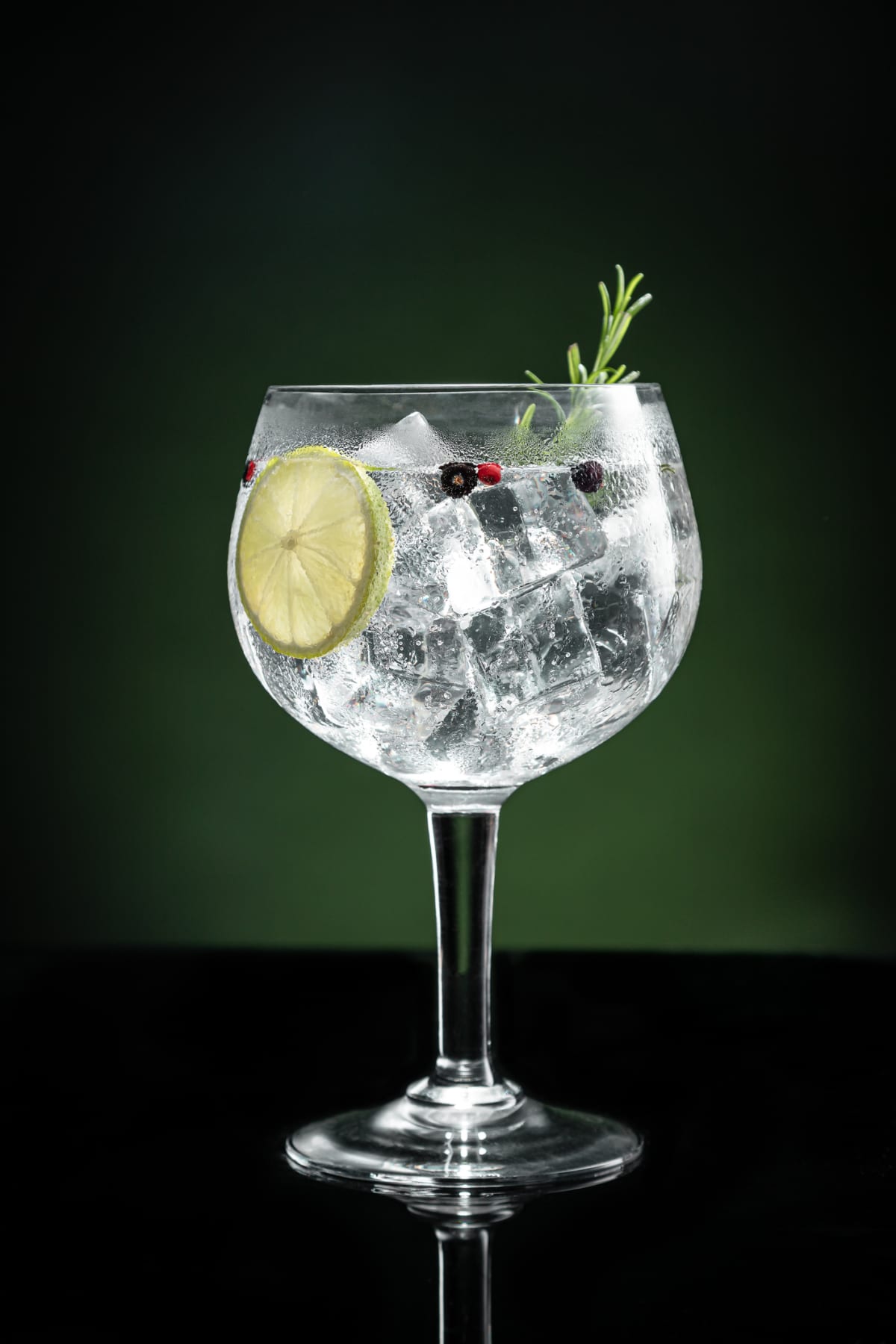 Cold glass of gin tonic with ice and lime, berry, and green garnishes