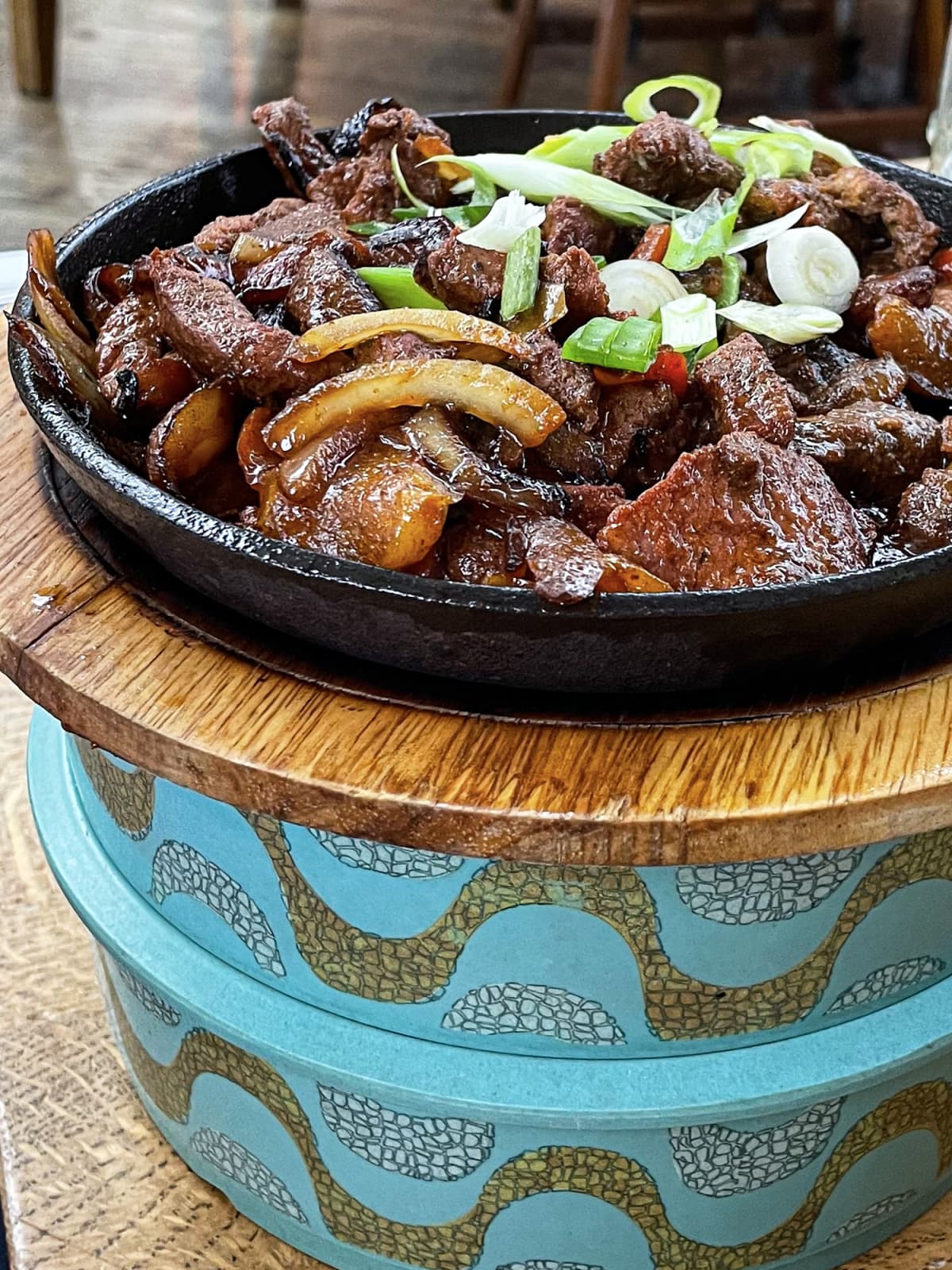 Cast iron pan on circular wood chopping board containing sizzling strips of grilled steak with stripped peppers and onions