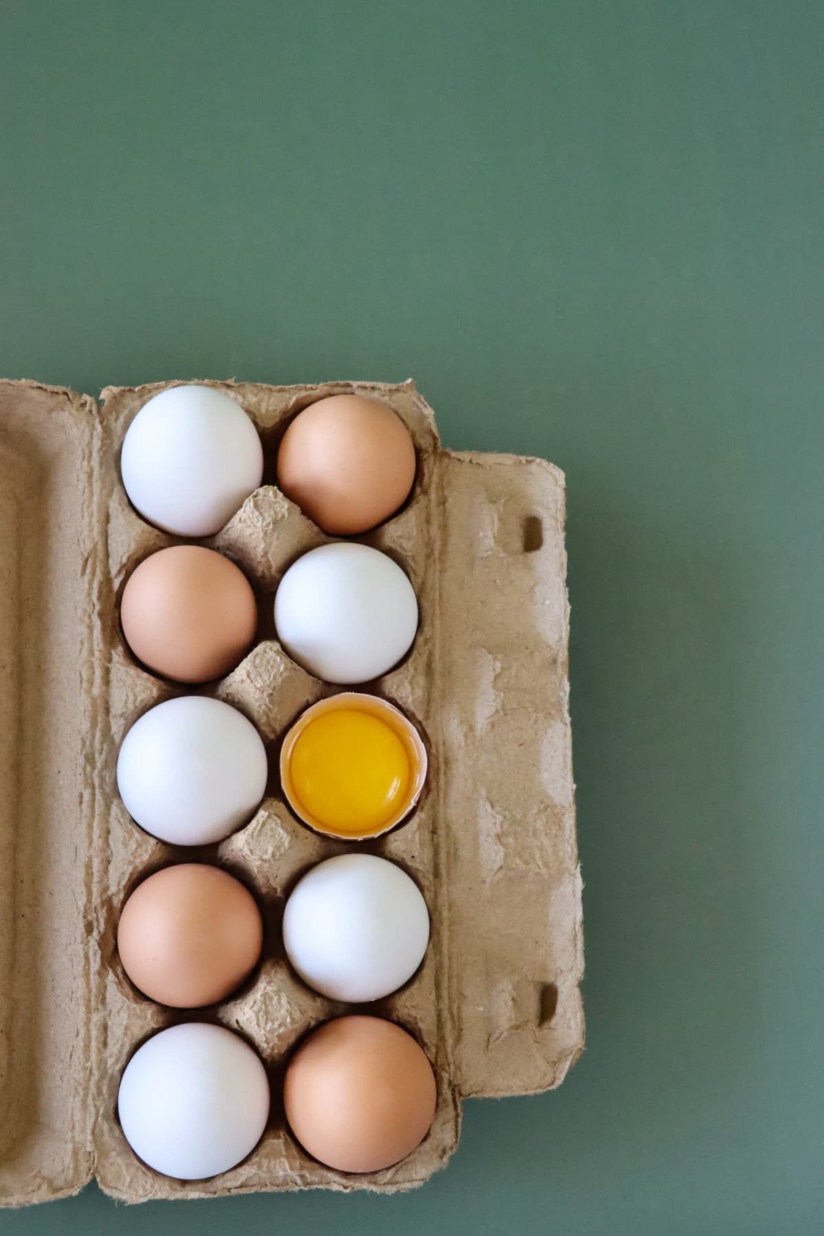 Stock photo showing elevated view of batch of ten mixed white and brown coloured eggs in open disposable cardboard egg box on green background.
